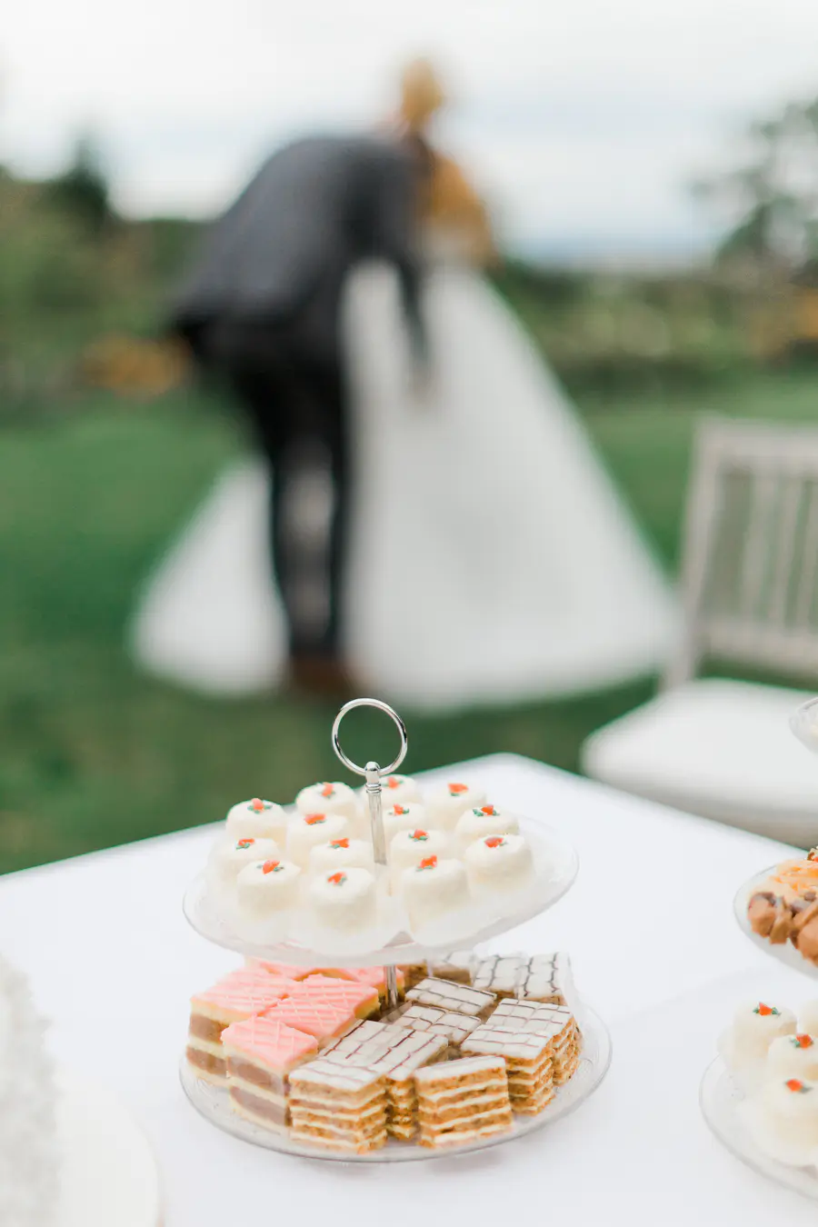 A close-up of a two-tiered dessert stand displaying various petit fours on a white tablecloth. In the blurred background, a bride in a white dress and a groom in a dark