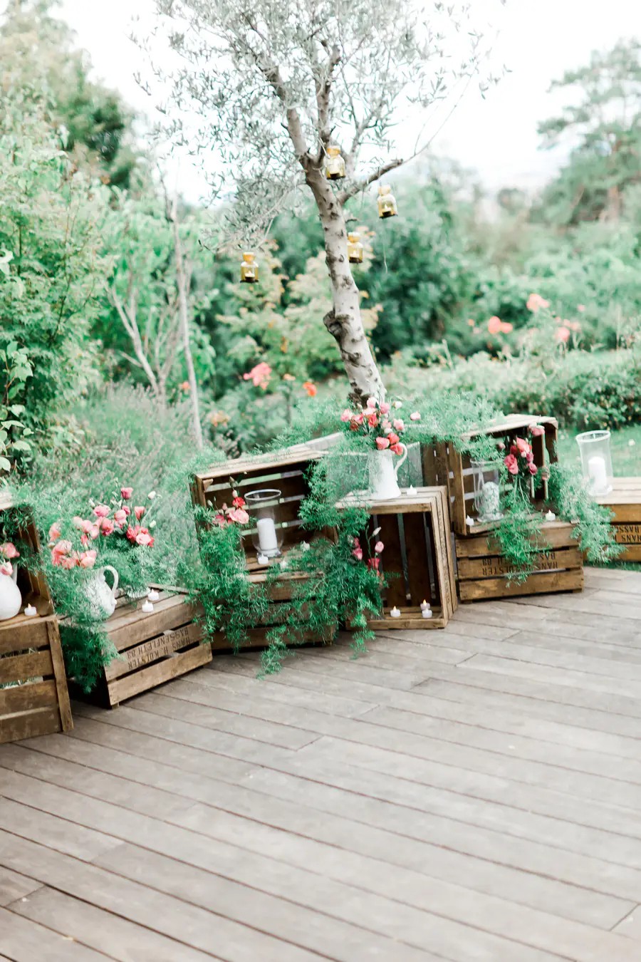 A rustic outdoor event setup with wooden crates arranged on a deck, adorned with pink flowers in white pitchers, trailing green foliage, and small candles. An olive tree with hanging yellow lanterns stands behind, surrounded by lush garden greenery.