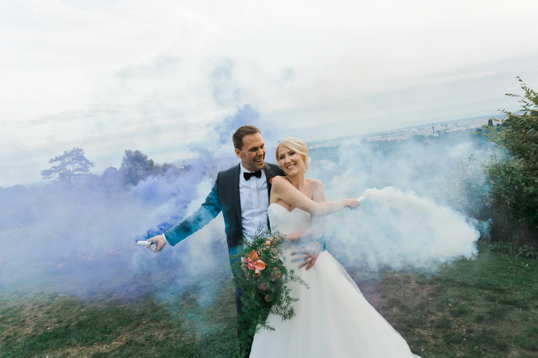 Weingut Reisenberg story: A smiling bride and groom in formal wedding attire stand outdoors, embracing and holding colorful smoke flares that billow blue and white smoke around them. A distant city skyline is visible in the background under a cloudy sky.