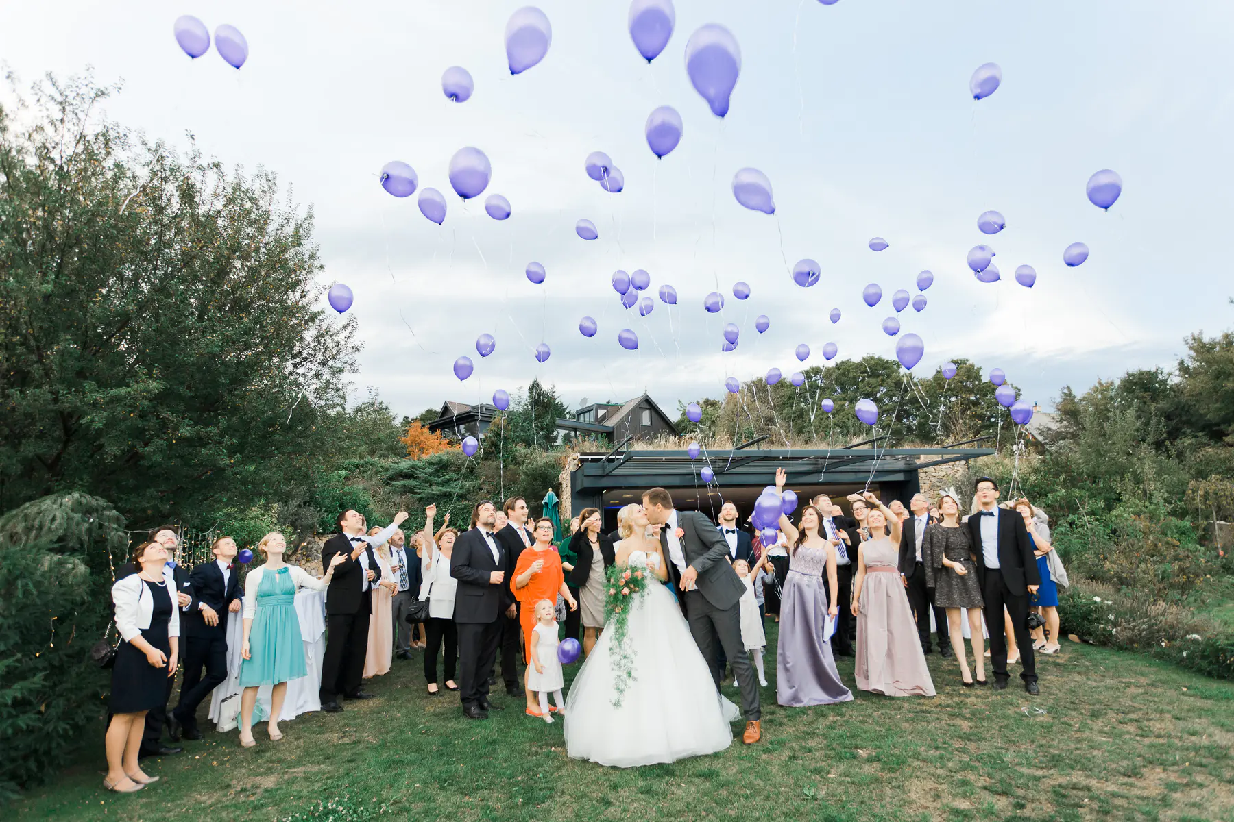 A newlywed couple kisses in the foreground, surrounded by wedding guests who look up as dozens of purple balloons with white ribbons float into the cloudy sky. The outdoor scene features green grass, lush trees,