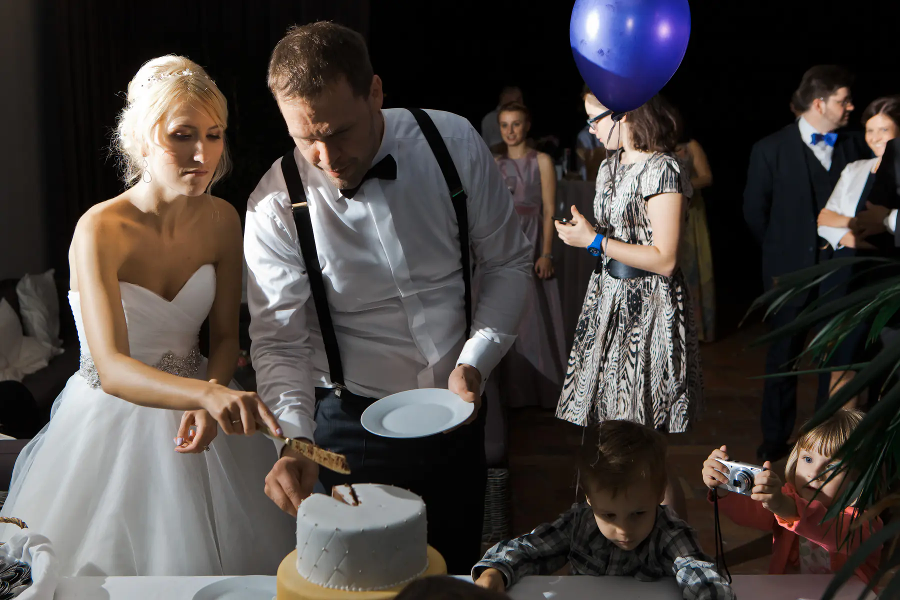 A blonde bride in a white strapless gown and a groom in a white shirt, bow tie, and suspenders cut a wedding cake together. They both look down at the cake, as guests socialize in the dimly lit background. Two young children watch from the foreground, one girl holding a small camera.