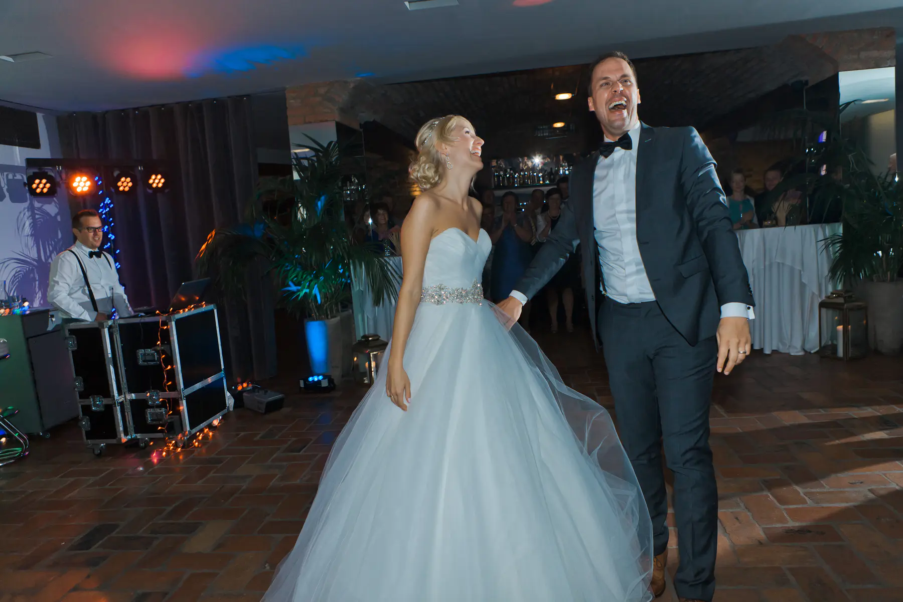A bride and groom are laughing exuberantly and walking hand-in-hand across a tiled dance floor. The bride wears a white strapless gown with a sparkling belt.Marek and Klara photography
