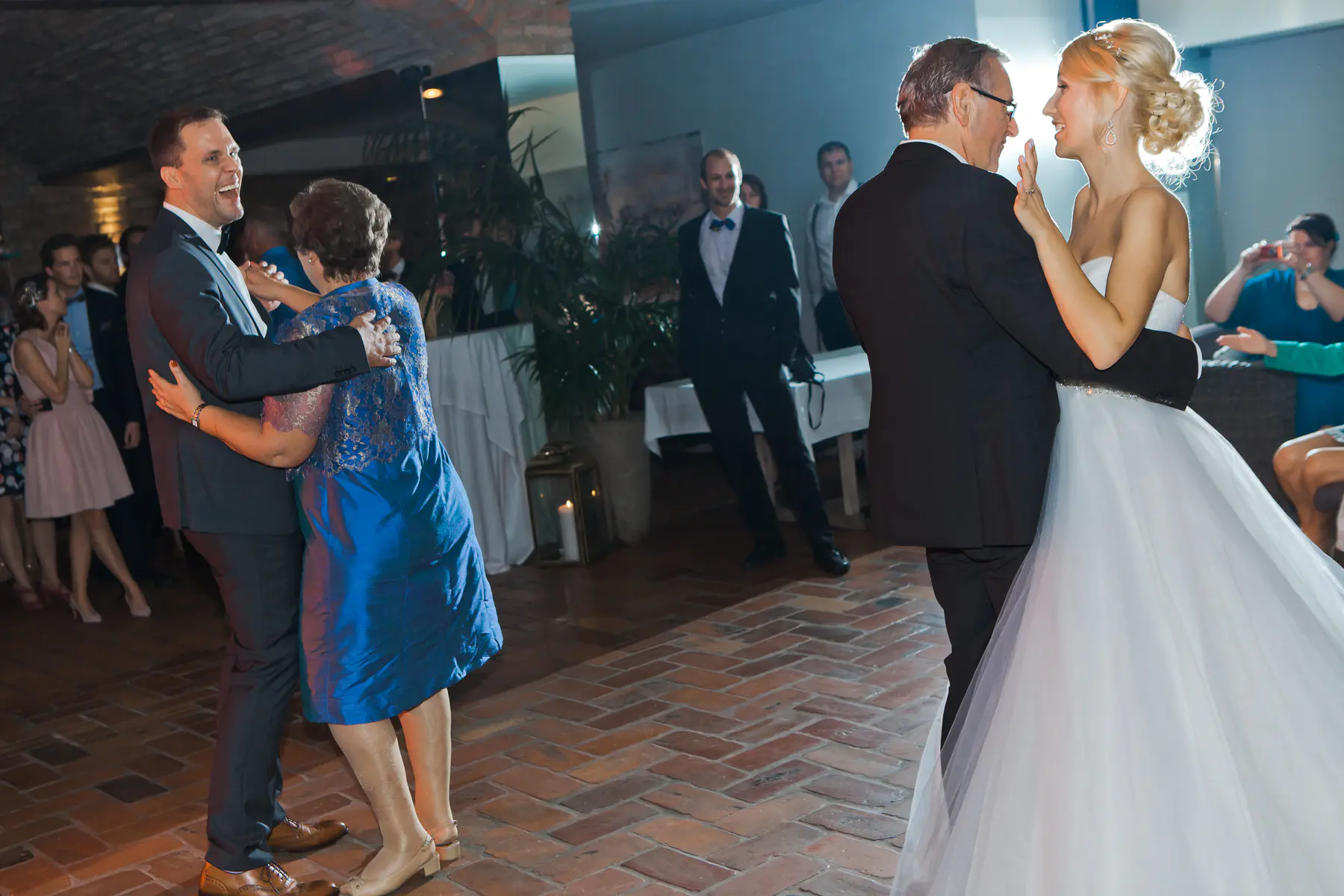 A joyful wedding reception shows a smiling bride in a white gown dancing with an older man on the right, while on the left, a smiling groom in a dark suit dances with an older woman.Weingut Reisenberg in Vienna