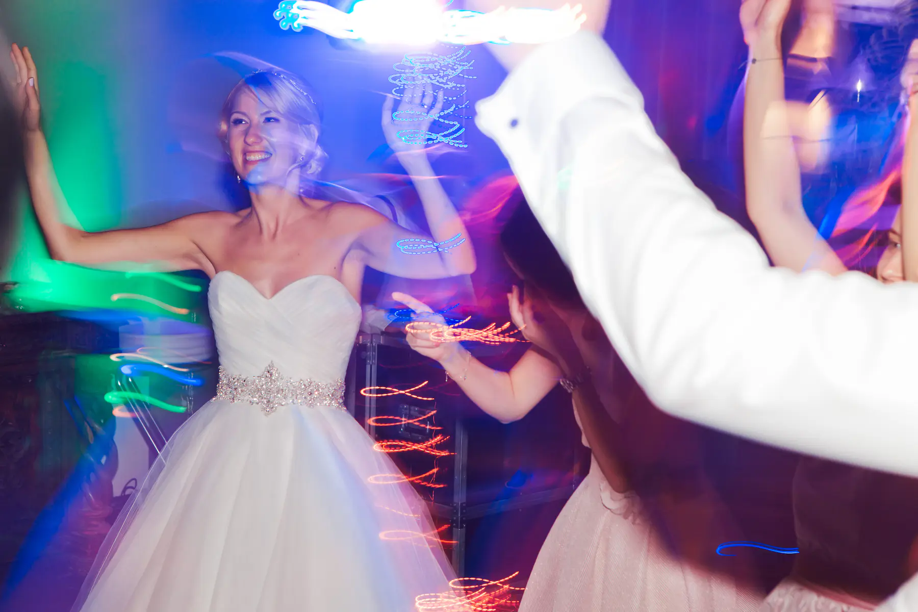 A bride in a white strapless wedding gown with a sparkly belt smiles and dances with her arms raised, surrounded by other blurred figures and streaks of colorful motion-blurred lights in blue,