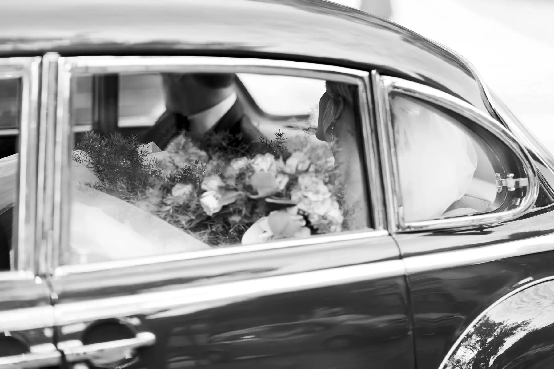 A black and white close-up shot shows the reflection of a bride and groom in the window of a vintage car. The bride is on the right, her face turned away, wearing a veil. The groom is on the left, wearing a suit. A bouquet of flowers sits between them.