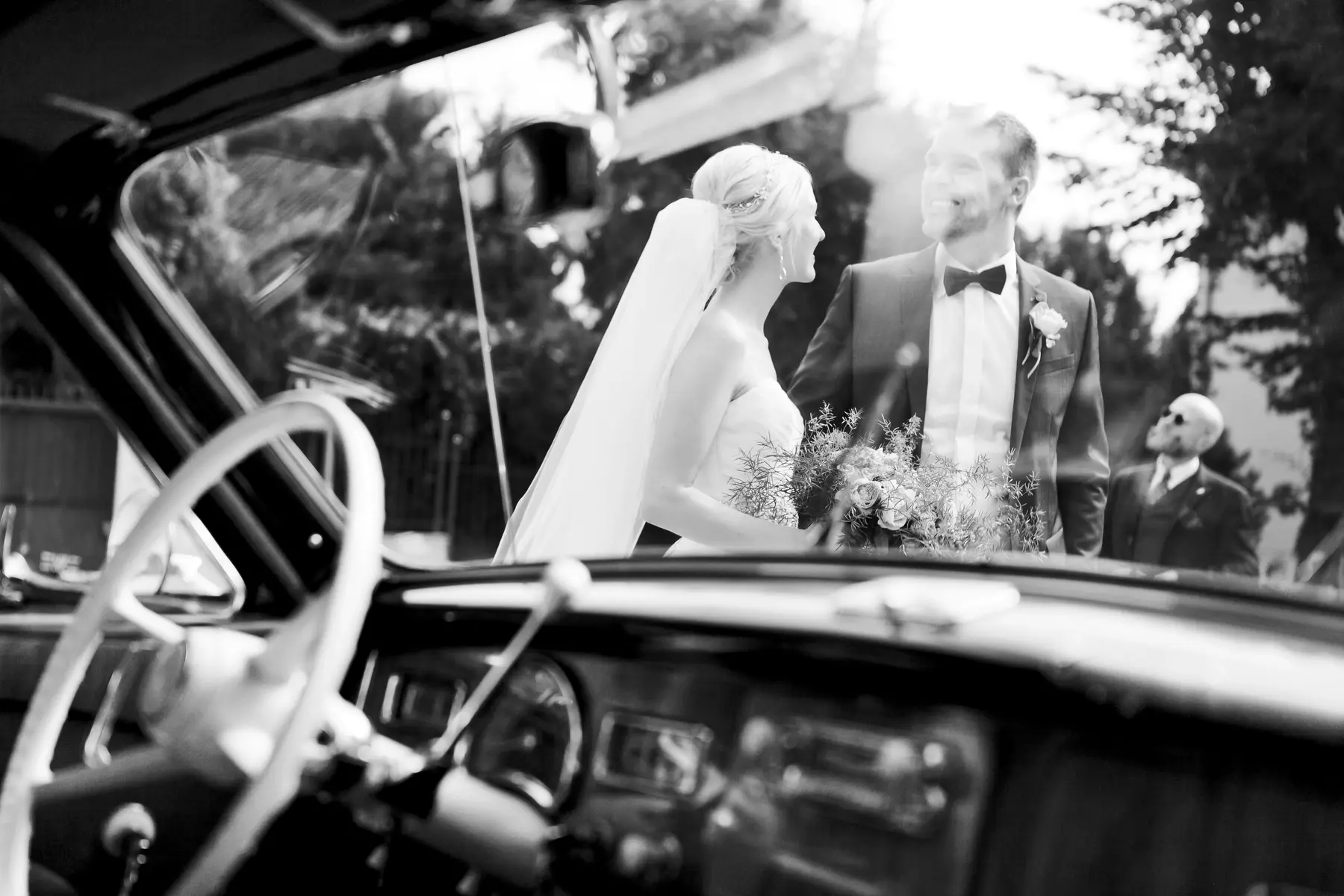 A black and white shot captures a newlywed couple standing next to a vintage car. The bride, on the left, wears a strapless gown and a veil, while the groom, on the right, is in a suit with a bow tie. They are holding a bouquet of flowers. The car's interior, including the steering wheel and dashboard, is visible in the foreground.