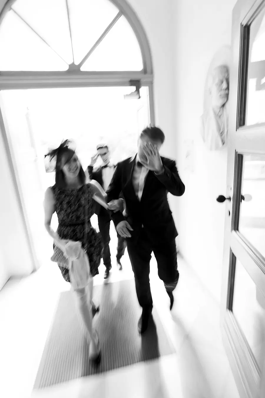A black and white, motion-blurred image shows a group of people in formal wear walking through an arched doorway. The woman in the foreground wears a dark, patterned dress and a hat, holding a light-colored bag. A man in a suit walks beside her, covering his face with his hand. Two other men in suits are visible behind them.