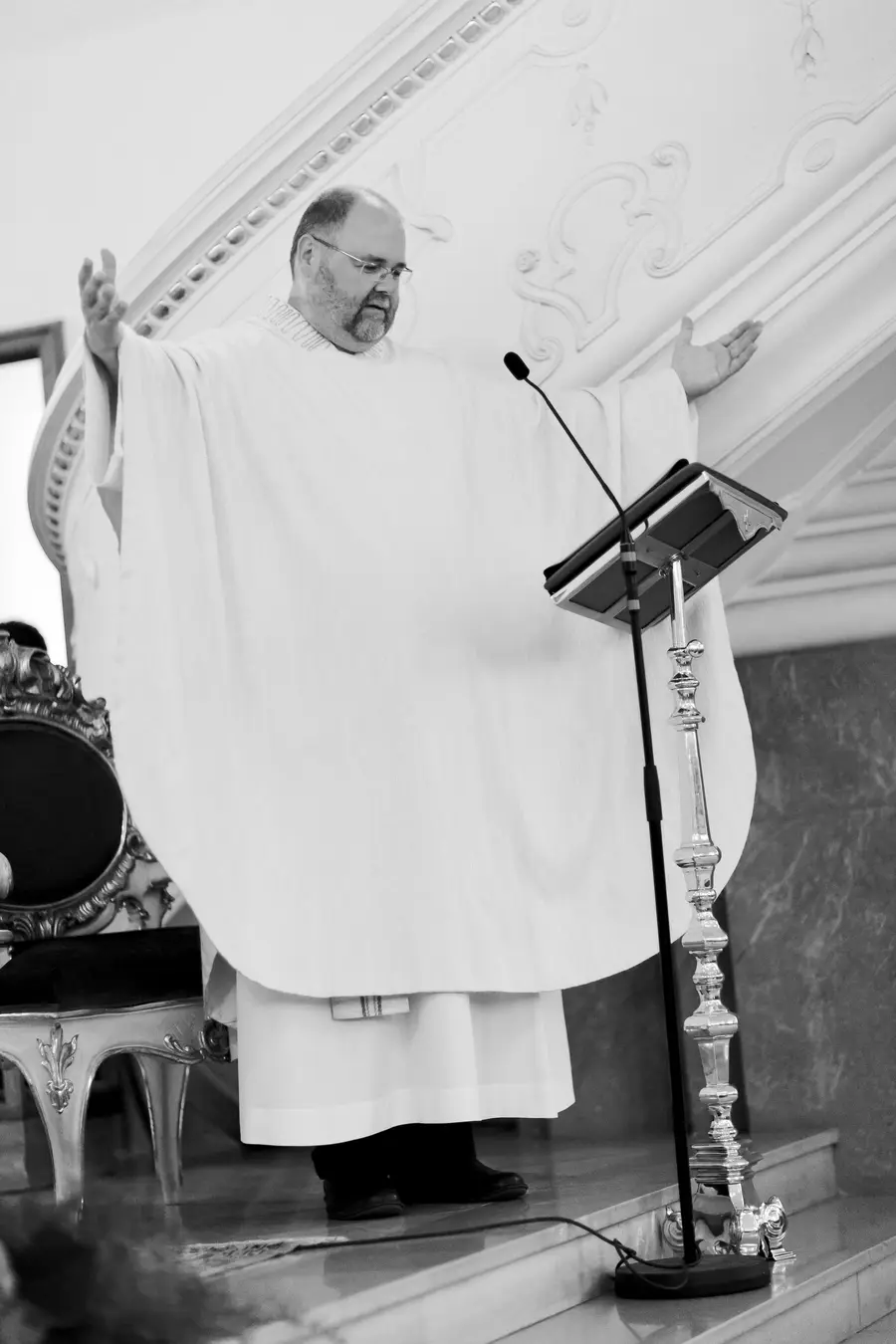 A man wearing a white liturgical robe stands with his arms outstretched and speaks into a microphone on a lectern in a church.