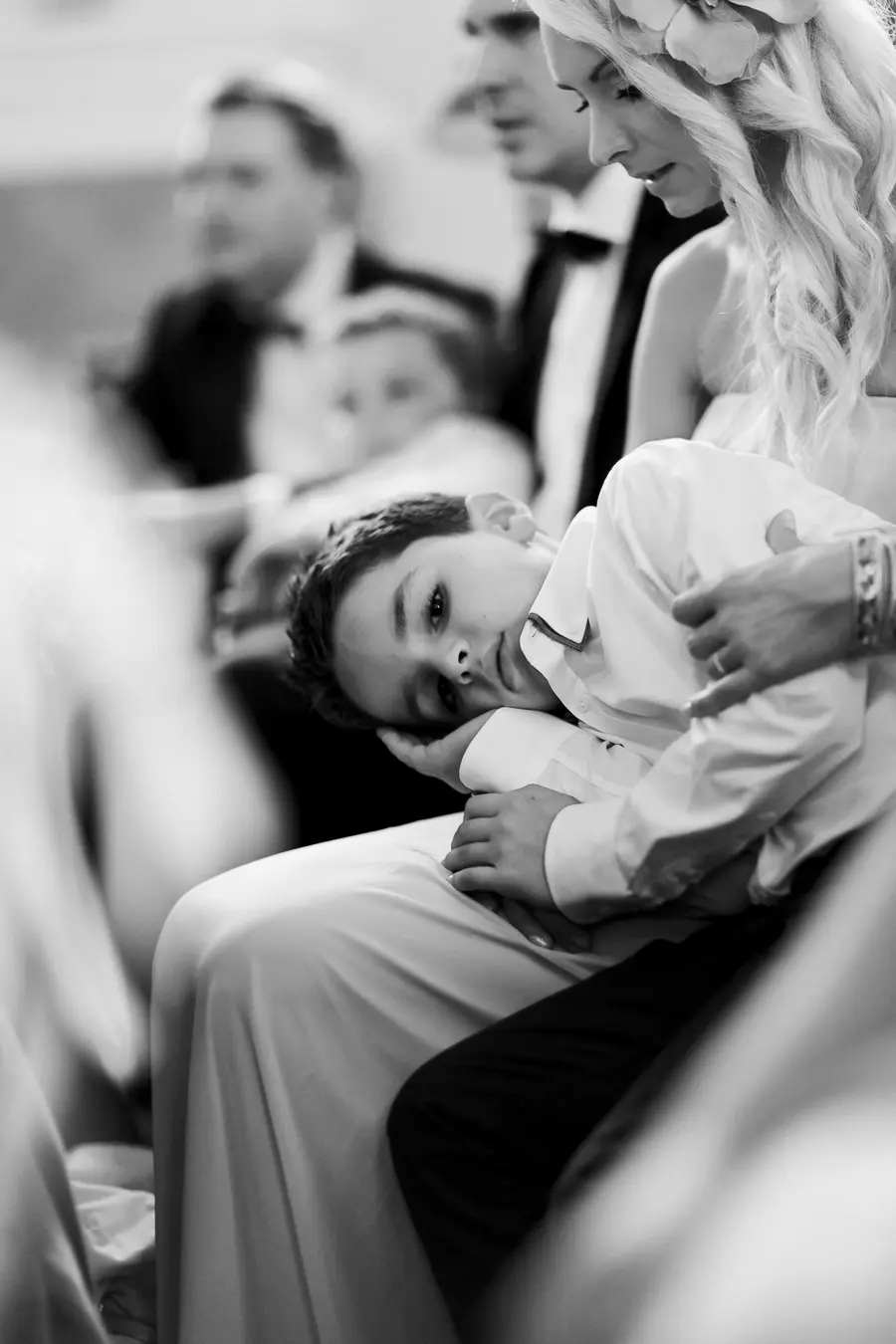 A young boy in a white shirt rests his head on his arm, looking at the camera. He is sitting on a woman's lap. In the background, several people in formal attire are visible.