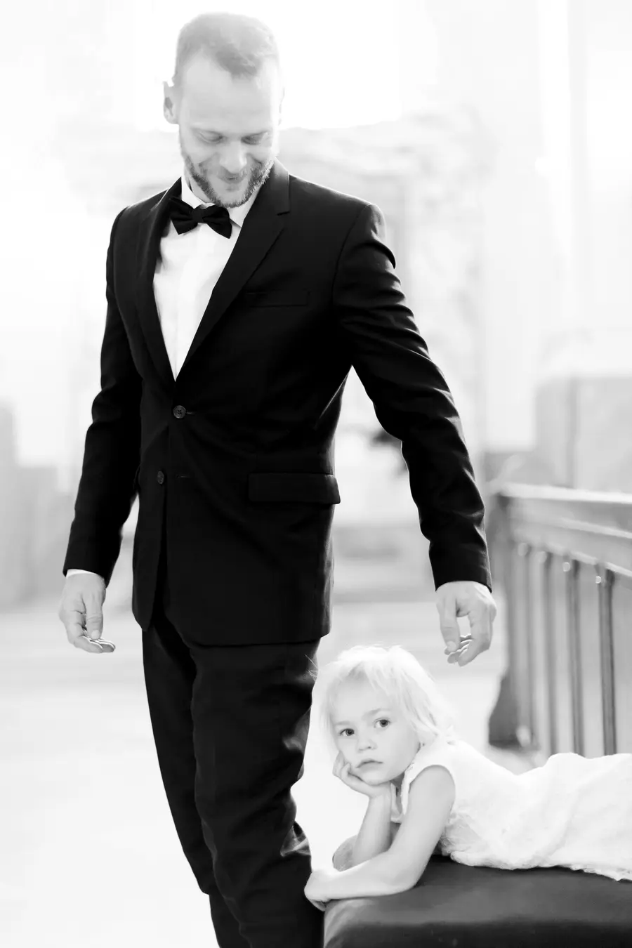 A man in a black tuxedo and bow tie looks down at a young girl in a white dress who is resting her chin on her hand.