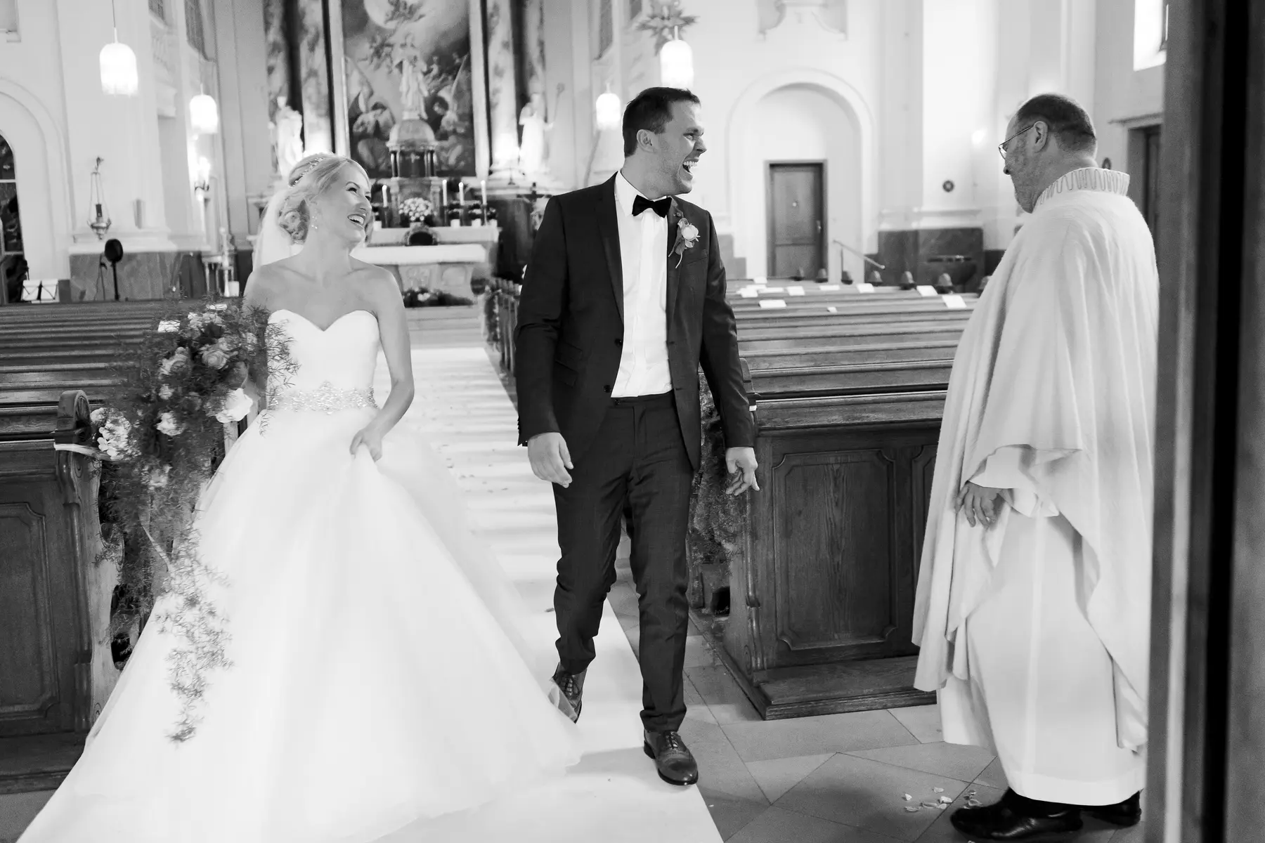 A bride and groom walk down the aisle in a church, laughing, as a priest stands to the side.Photojournalistic wedding photography Vienna