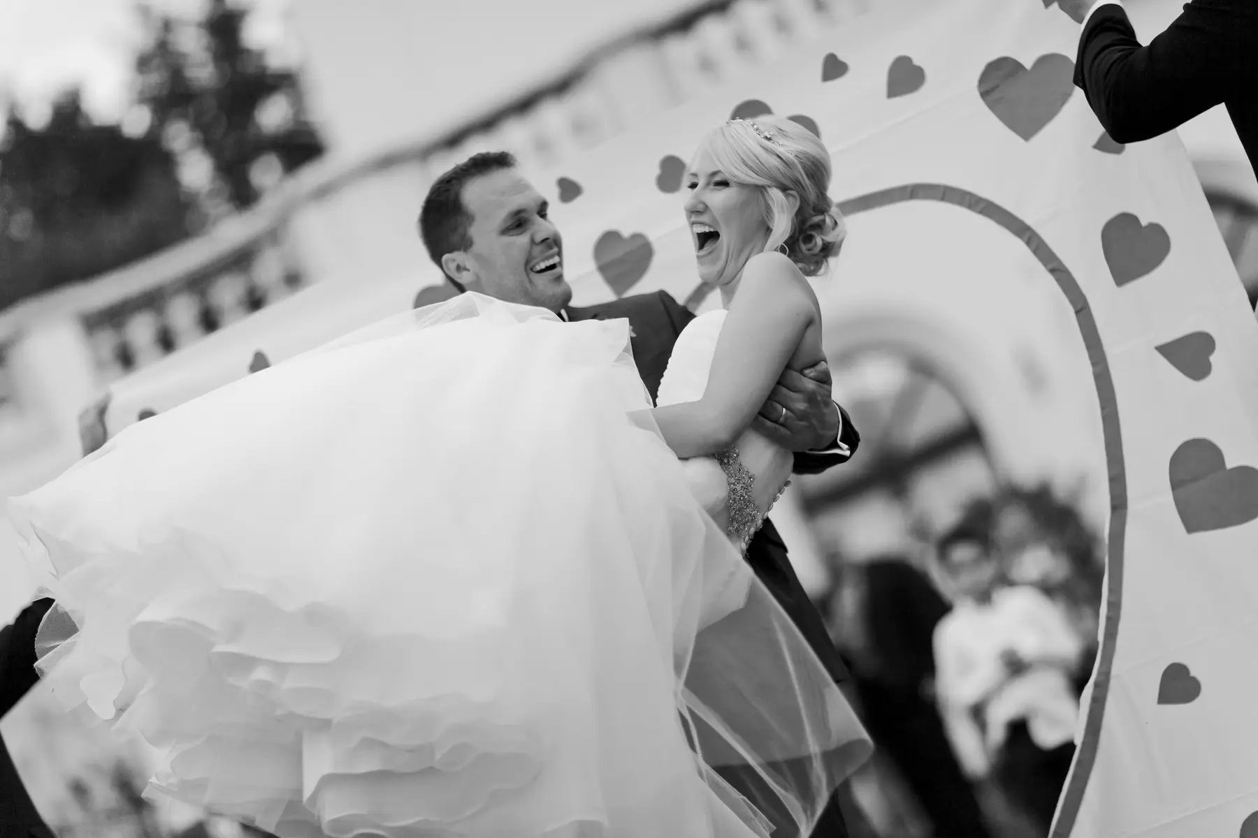 A groom joyfully lifts his bride in a black and white, medium shot. The bride, wearing a strapless wedding dress and a tiara, laughs with her mouth wide open as her groom spins her around. The groom, in a suit, also smiles broadly. They are in front of a large banner decorated with hearts and a circular arch, with blurred figures in the background.