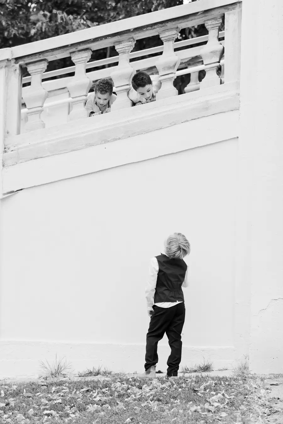 A black and white photograph shows three young boys. Two boys are peeking over a white ornate railing from above, with their heads down. Below them, a third boy stands with his back to the camera, looking up at the other two. He is wearing a vest and dark pants.