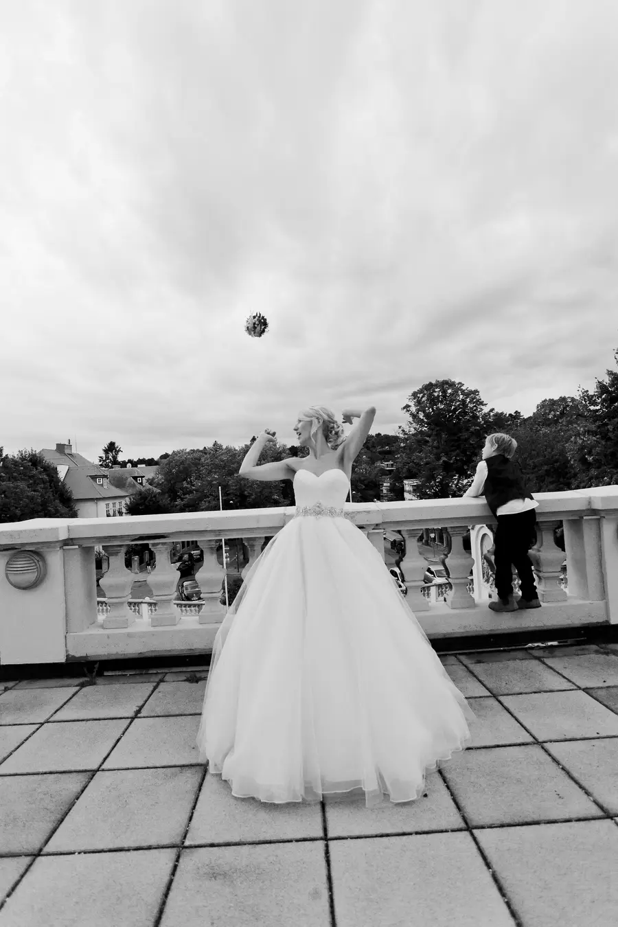 A bride in a full white wedding dress throws her bouquet into the air while a young boy in a vest watches from a balcony.