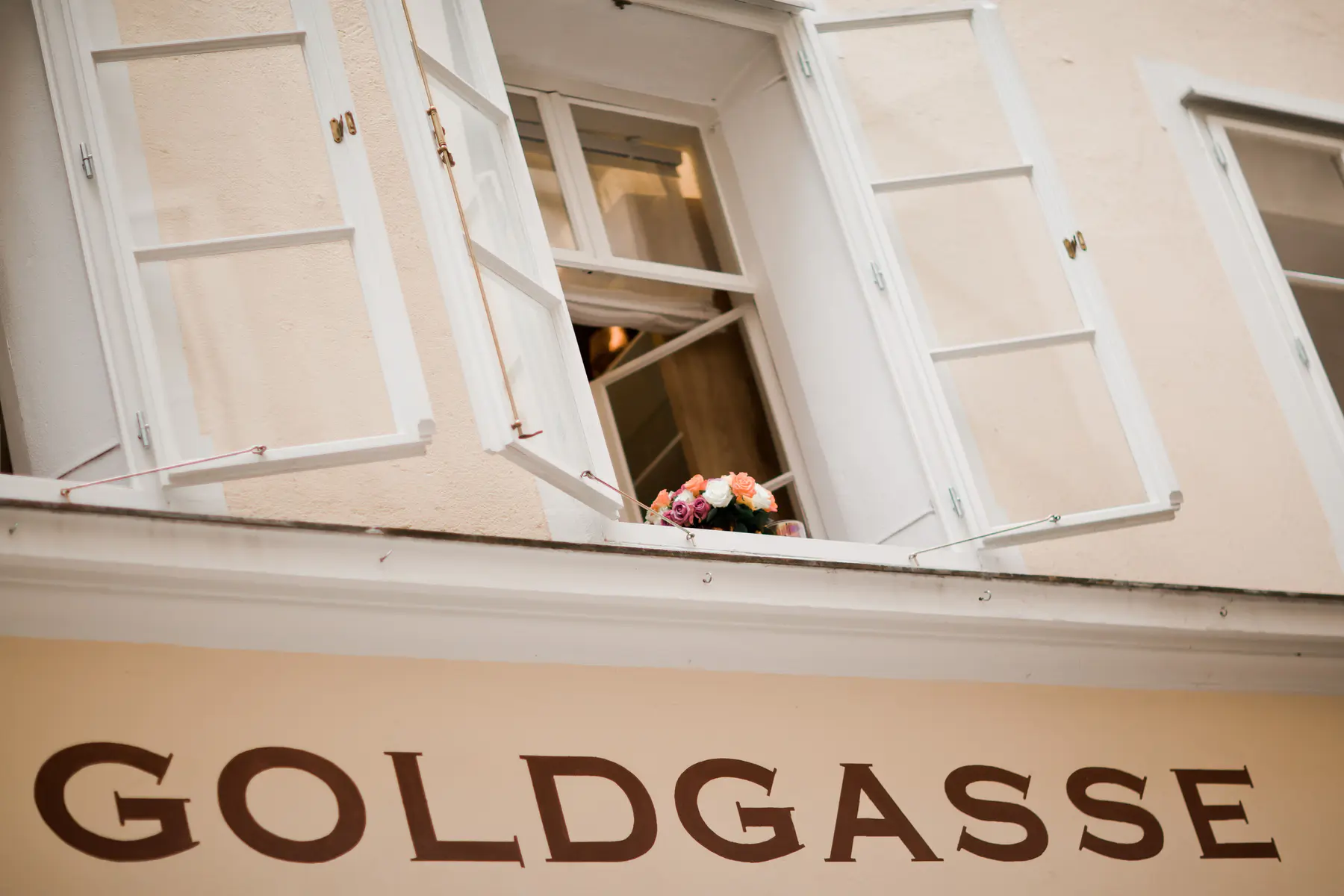 A light-colored building facade with two open white-framed windows. A small bouquet of pink, orange, and white flowers sits on the windowsill. Below the windows, a brown sign prominently displays the