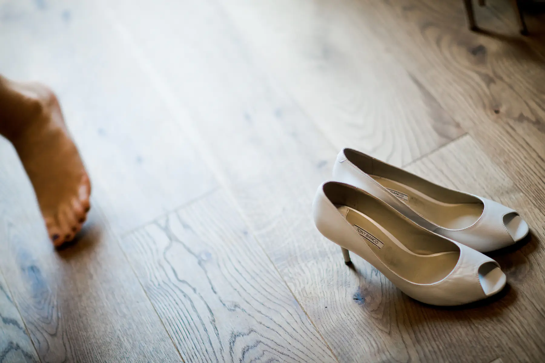 A pair of cream-colored, peep-toe Tony Bianco high heels sits on a light wooden floor. A blurry bare foot with dark toenail polish is visible in the background to the left.