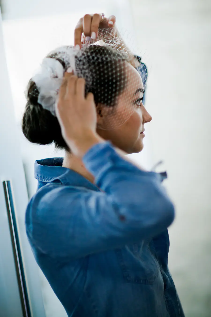 A bride with dark hair in an updo, wearing a blue collared shirt, adjusts a white birdcage veil with floral accents, seen in profile.