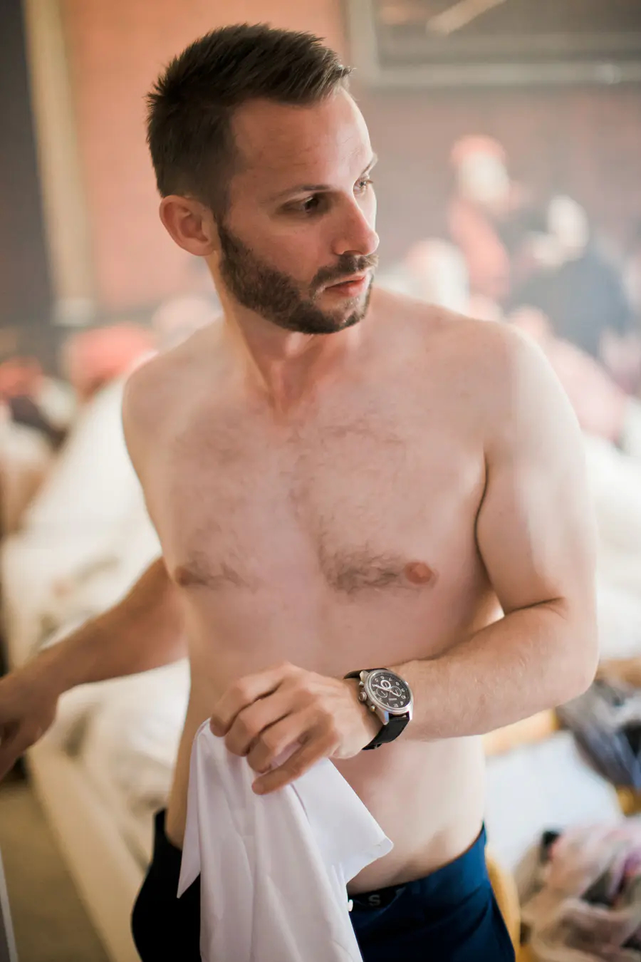 A shirtless groom with a beard and a watch holds a white shirt, looking to the side.Documentary Wedding Photography Salzburg