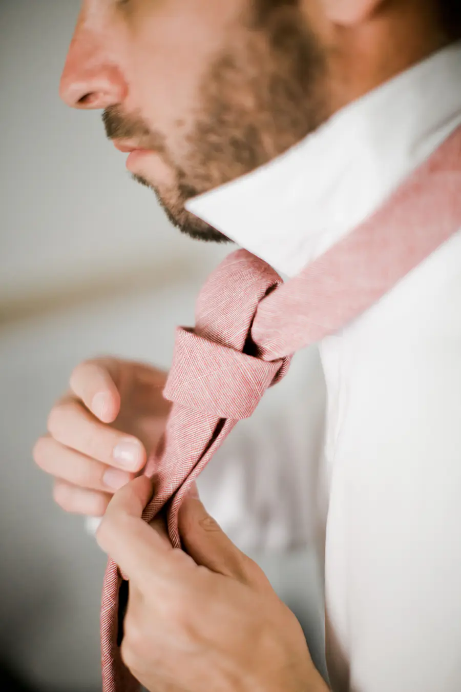Close-up of a groom adjusting a textured pink tie over a white shirt.