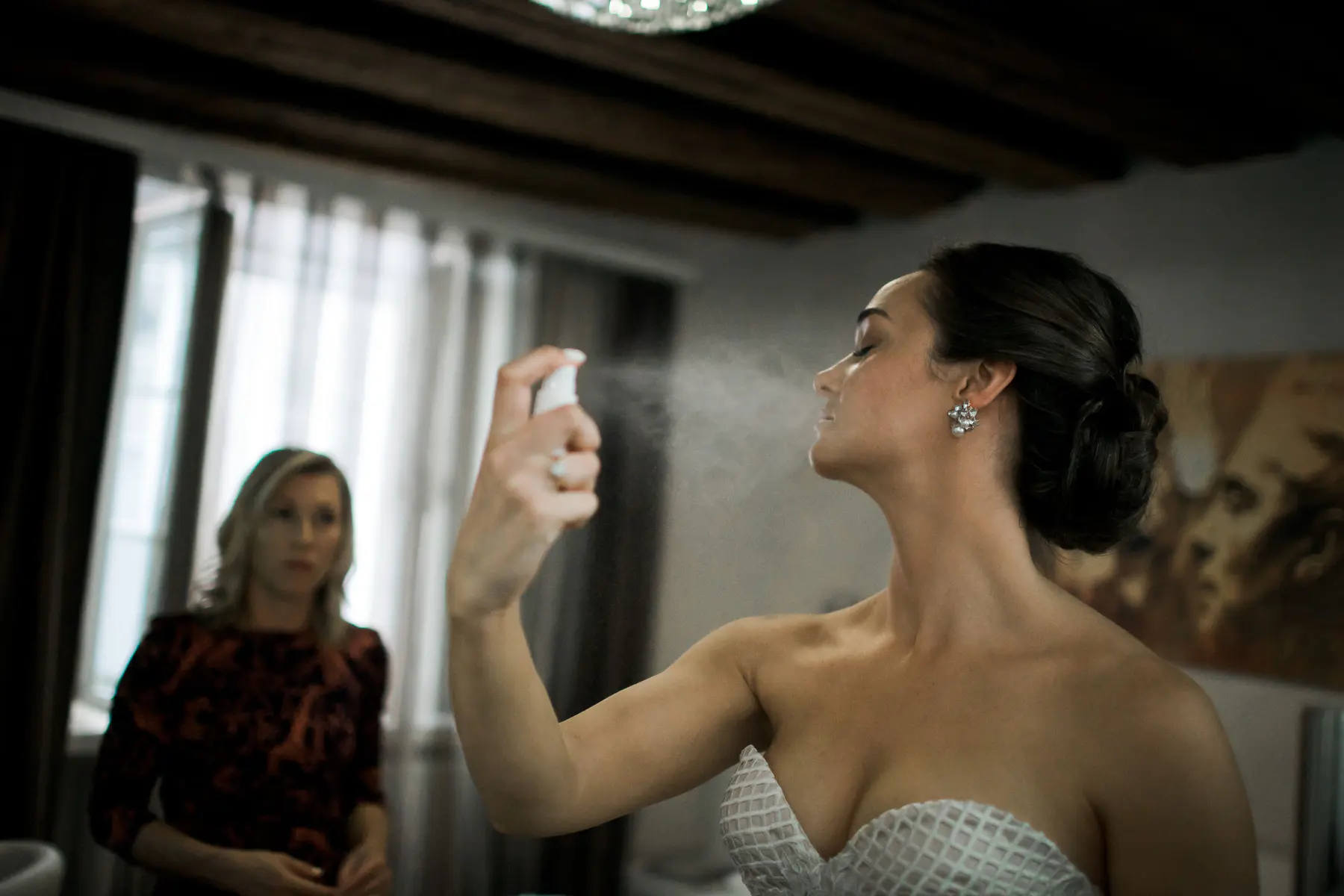  Hotel Goldgasse. A bride in a strapless white top sprays a fine mist onto her face from a white bottle, with a blurred woman observing in the background.