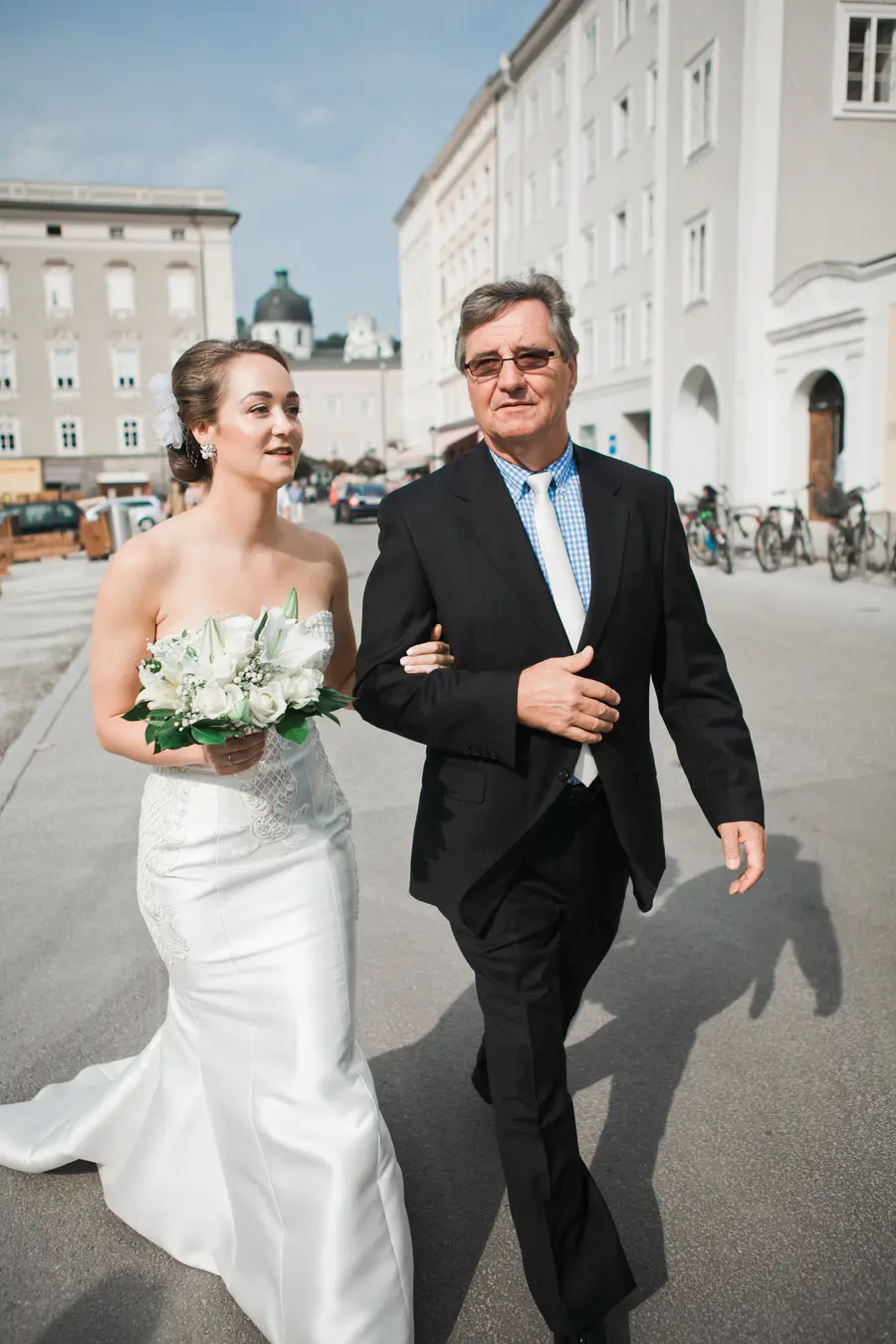 A bride in a strapless white gown holding a white floral bouquet walks arm-in-arm with an older man in a black suit down a sunny city street lined with historic European buildings.