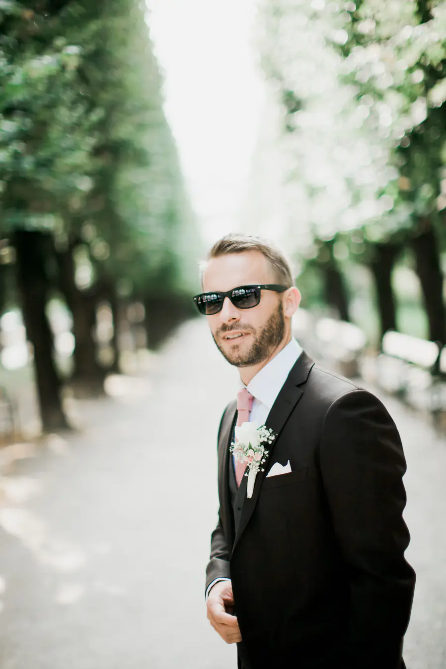 Groom in sunglasses smiles in tree-lined path.