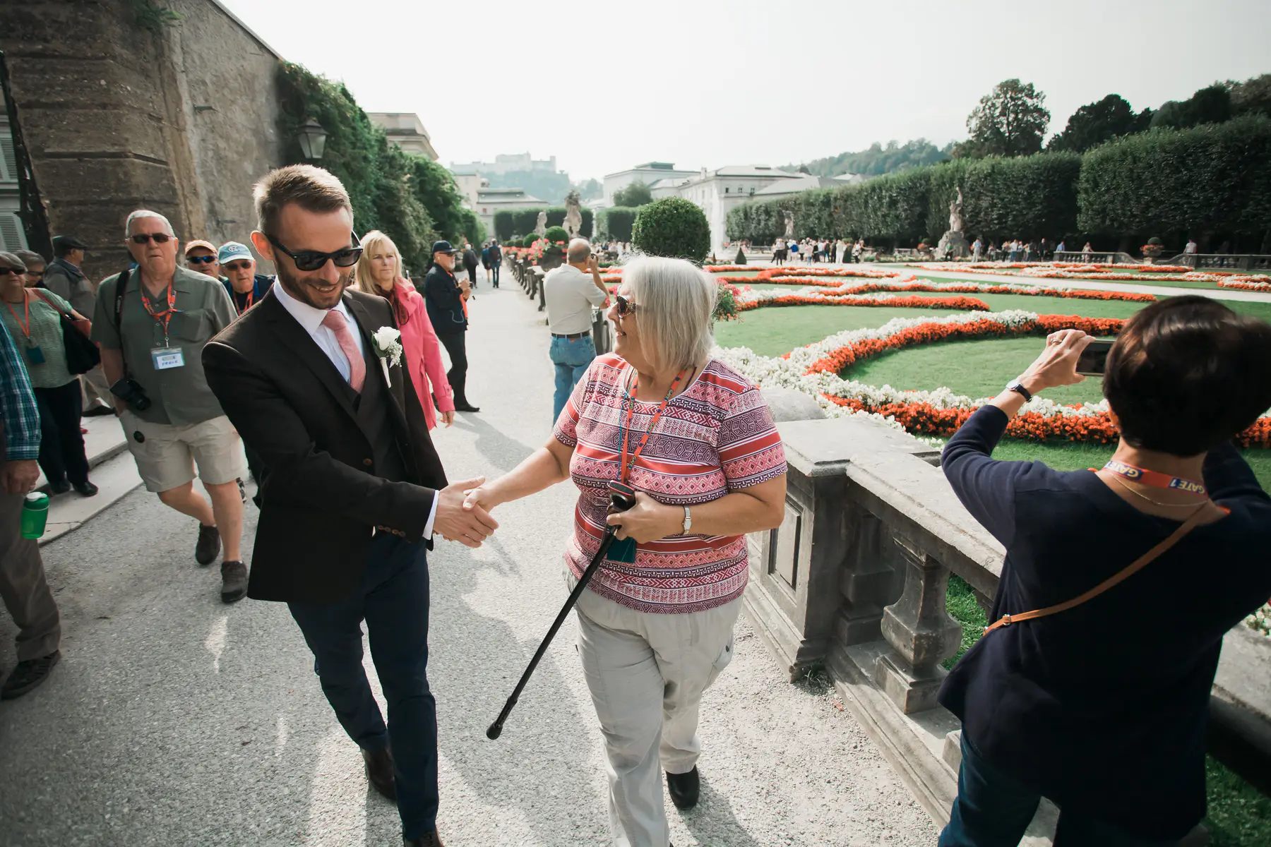 A groom in a dark suit with a boutonnière and pink tie shakes hands with an older woman holding a walking stick, both smiling. They are on a paved path in a formal garden with white and red flower beds to their right, and other people strolling or taking photos in the background.