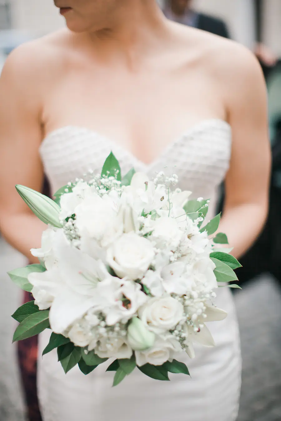 A bride in a white strapless dress holds a lush bouquet of white roses and lilies.