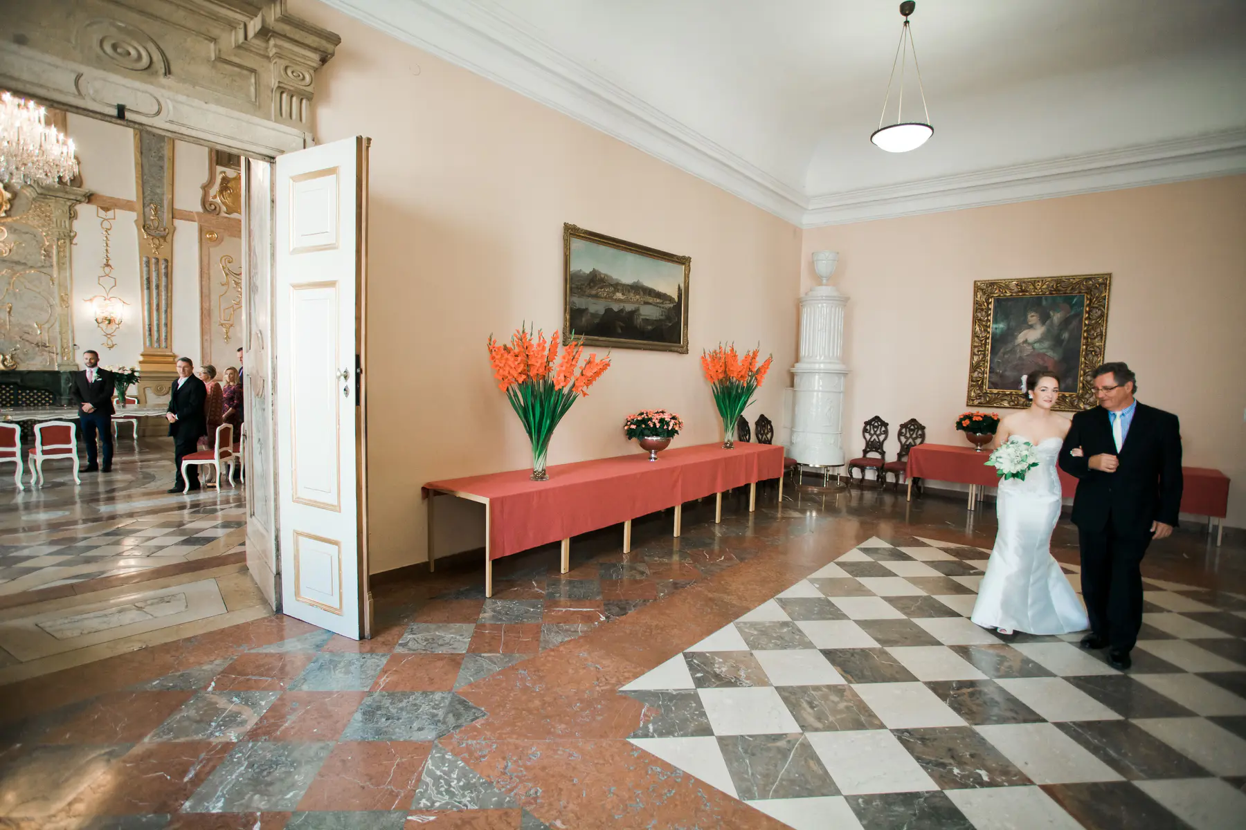 Mirabell Palace wedding photojournalism. A bride in a white gown walks down an aisle with her father, escorted by a man in a suit. The floor is a checkered pattern of black and white.
