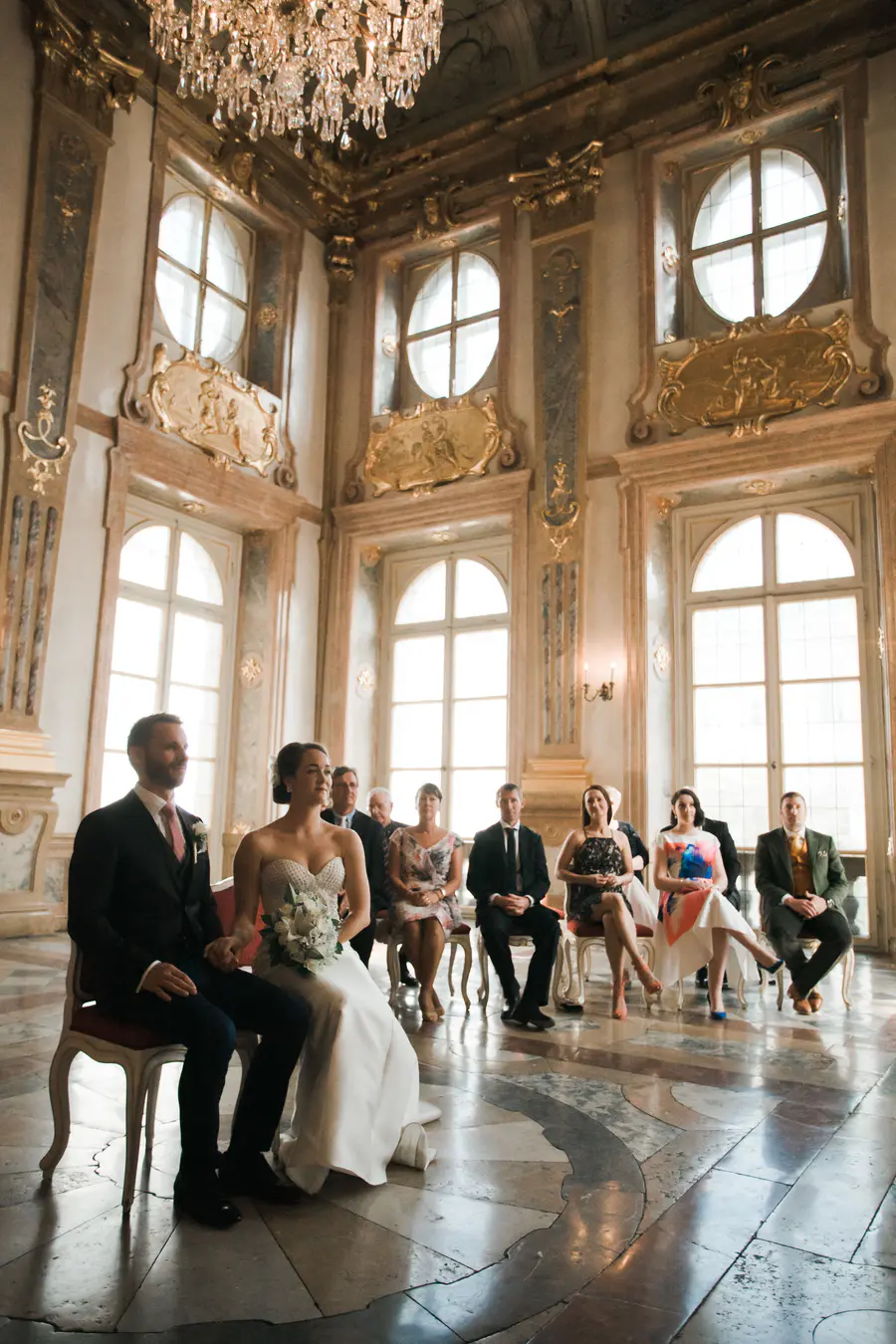 A wedding ceremony takes place in a grand, ornate room with high ceilings, tall windows, and golden architectural details. The bride and groom are seated in the foreground, facing right. The bride wears a strapless white wedding gown and holds a bouquet of white flowers. The groom is in a dark suit with a pink tie. Behind them, several guests are seated in chairs, also facing right. The room is lit by natural light streaming through the windows and a large crystal chandelier hanging from the ceiling.