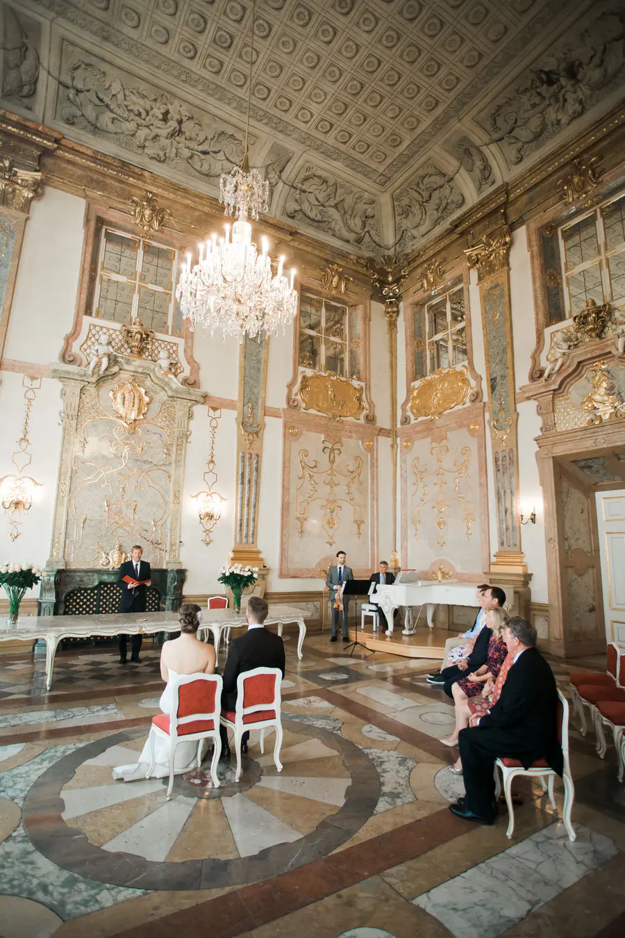 A wedding ceremony is taking place in a lavishly decorated hall with a grand chandelier. A couple sits facing forward, with an officiant to their left and musicians to their right.