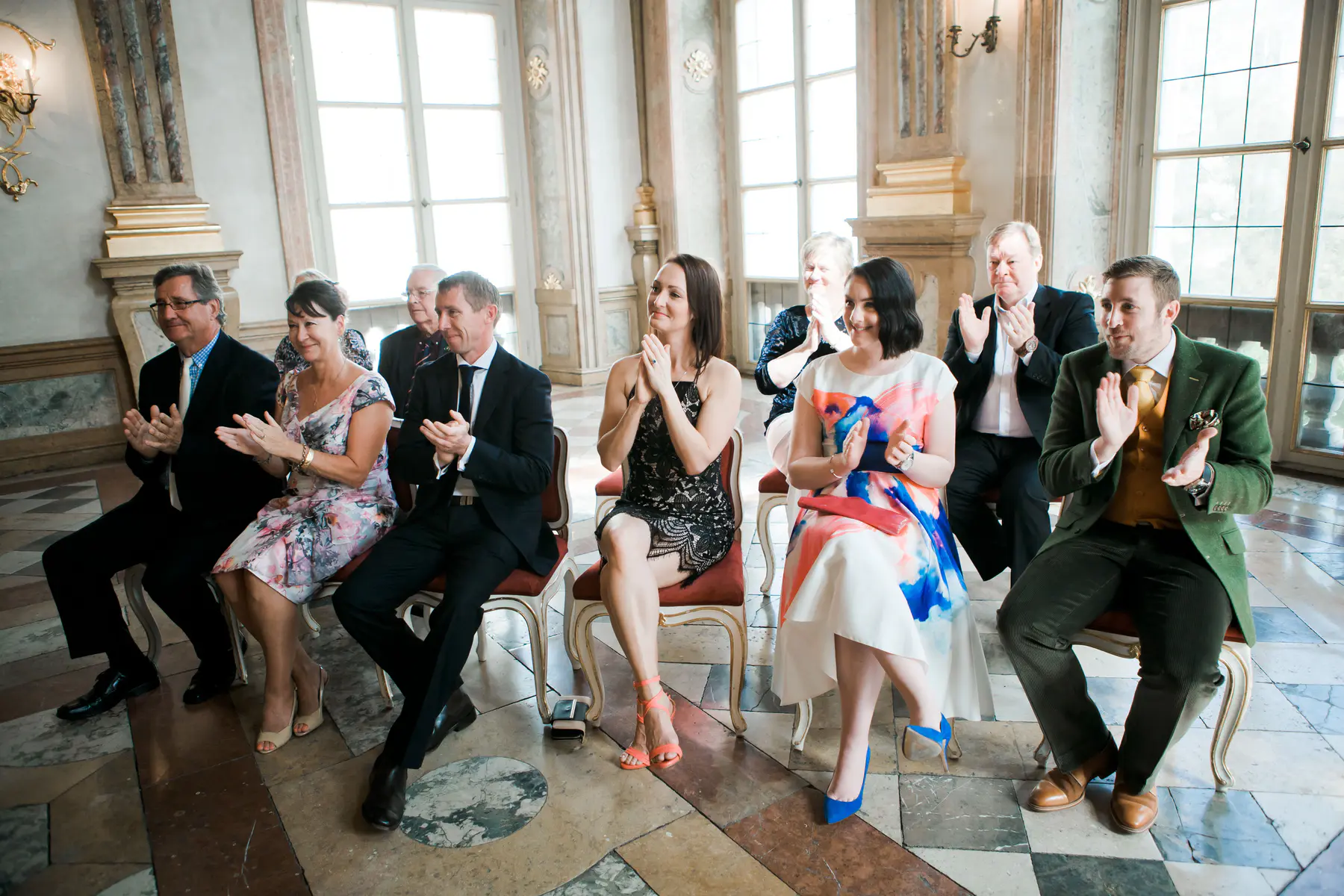 A group of people are seated and clapping in a room with large windows and ornate decor.
