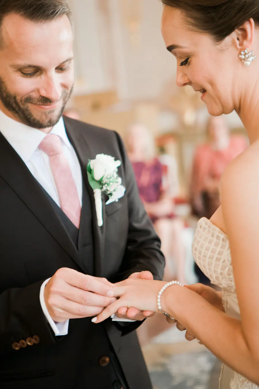 Wedding ring exchange in the Marble Hall Salzburg. A groom in a black suit and pink tie places a ring on the bride's finger during a wedding ceremony.