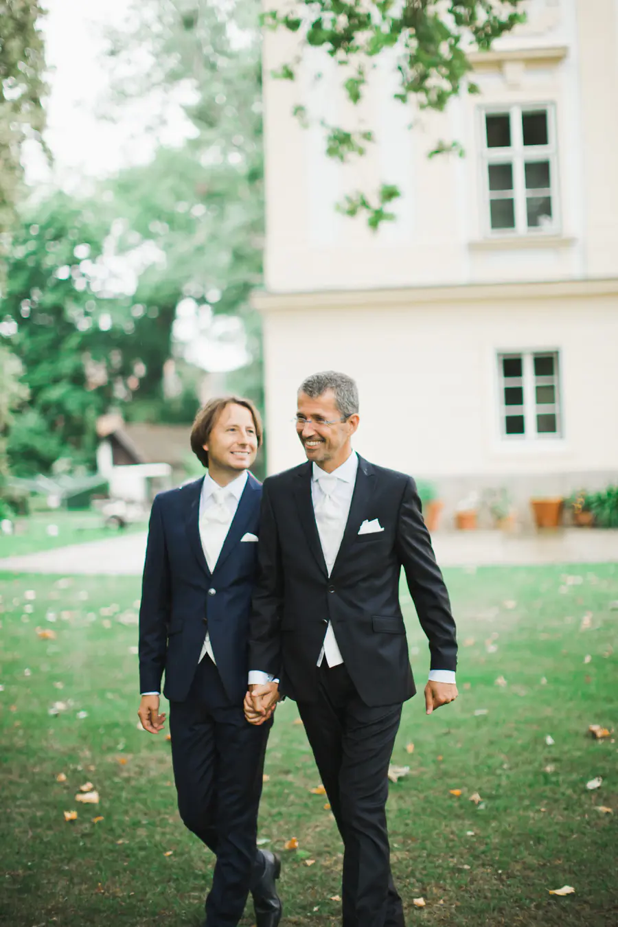 玛格丽滕城堡 (Schloss Margarethen am Moos) 的真实婚礼纪实。两位新郎在宏伟的城堡花园中手牵手漫步。下奥地利州的现代、非摆拍纪实摄影。捕捉动人瞬间与专属视觉语言。Marek & Klára 为每一份爱见证。