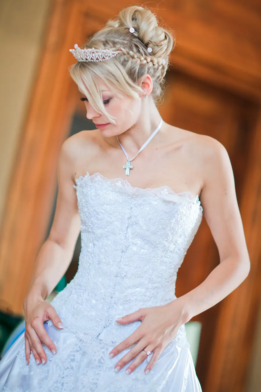 A bride with blonde hair styled in an updo with braids and adorned with a tiara and small flowers, wearing a white strapless wedding dress with intricate beadwork and a lace trim. She is also wearing a white ribbon necklace with a cross pendant and a diamond ring. She is looking down with her hands resting on her dress.
