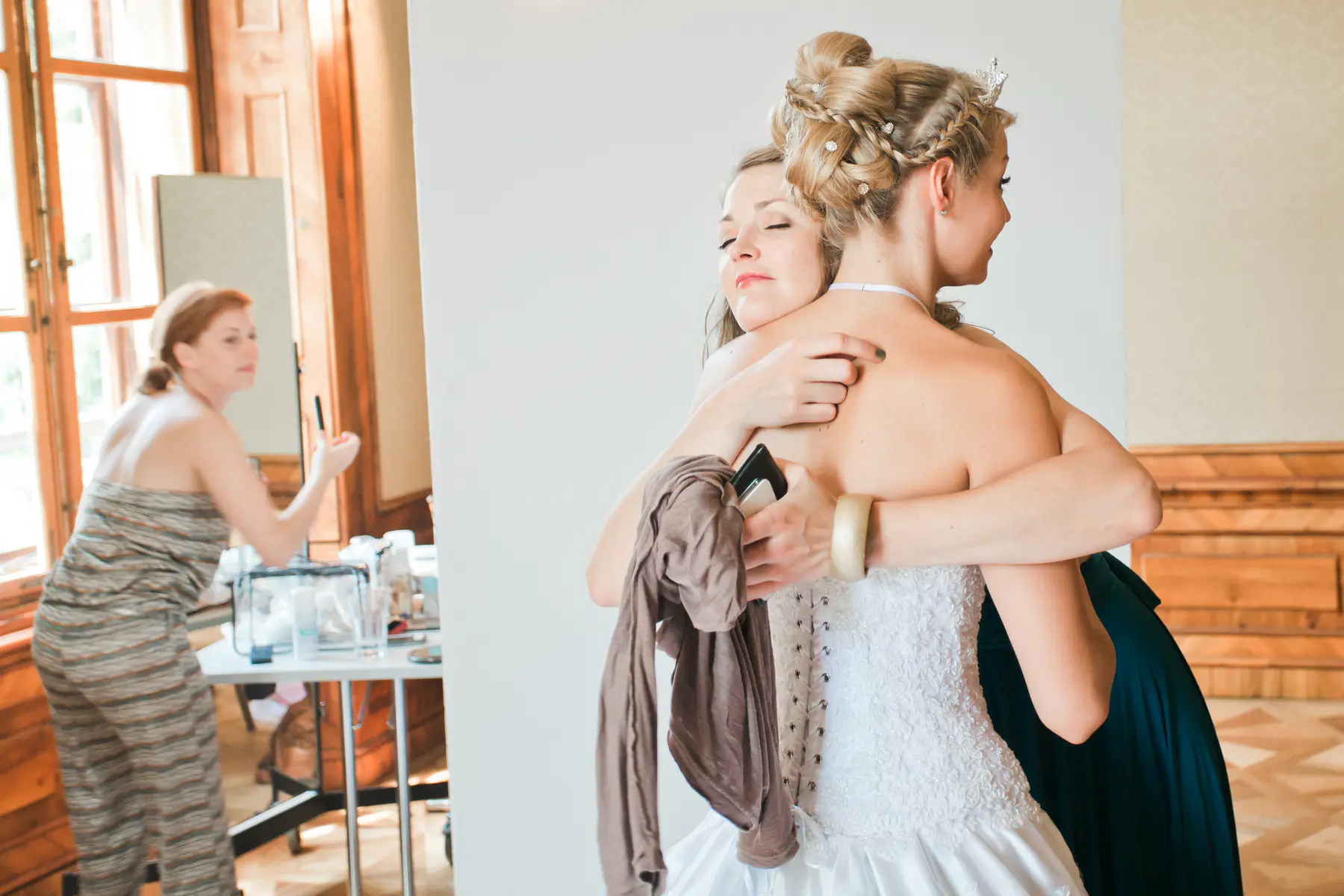 A bride with her hair in an elegant updo is being hugged by a woman in a white dress, with a third woman in the background attending to makeup.