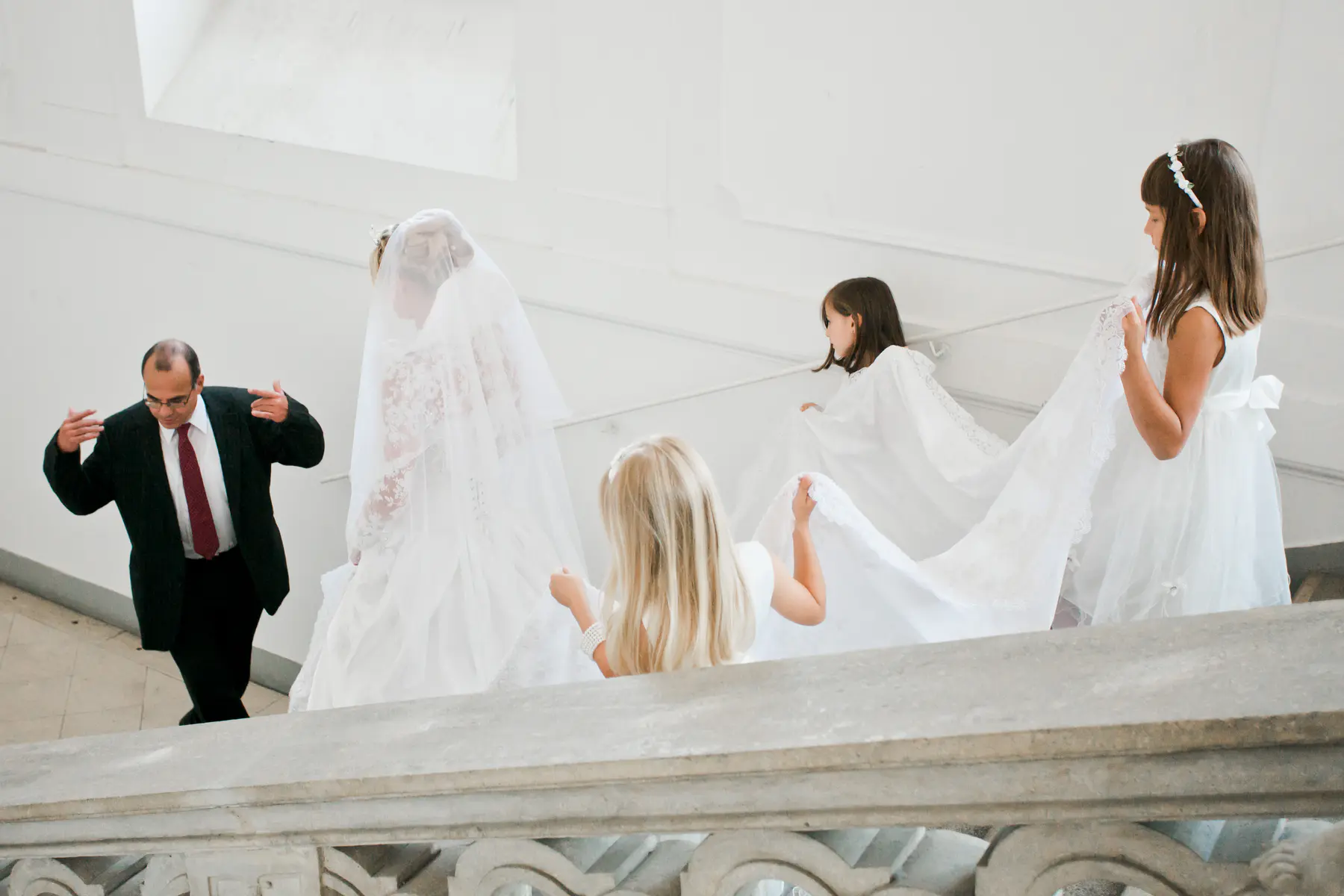 A father in a suit and tie helps hold the train of a bride's wedding dress as they walk up a white staircase. Two young girls in white dresses are also helping to carry the train.