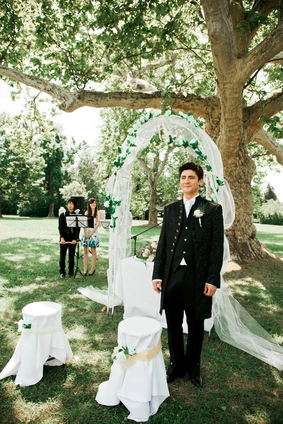 A groom in a black ornate tuxedo stands outside under a large tree. He is smiling as two musicians and a woman in a blue and white dress stand behind a decorated wedding arch in the background.