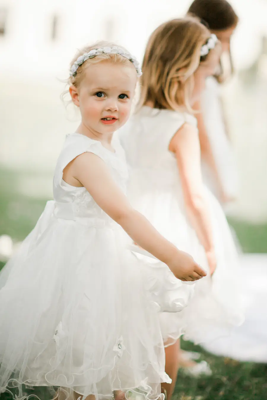 A young girl with a flower crown looks toward the camera, her blonde hair framing her face. She wears a white, ruffled dress and stands in front of two other girls also in white dresses.