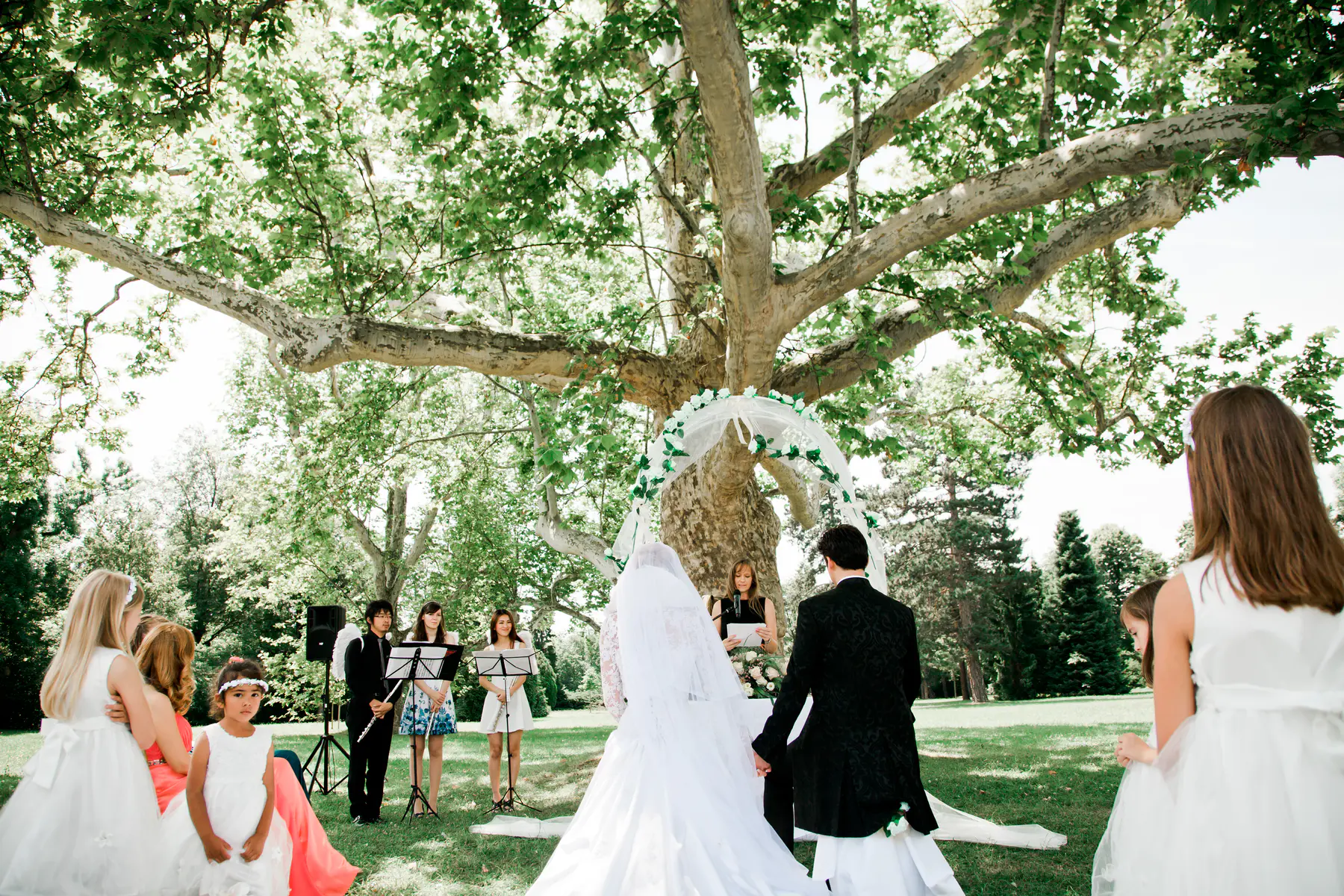 A wedding ceremony is taking place outdoors under a large, leafy tree. The bride and groom stand facing each other beneath a floral arch. A few bridesmaids and flower girls in white dresses are visible, along with musicians playing instruments.
