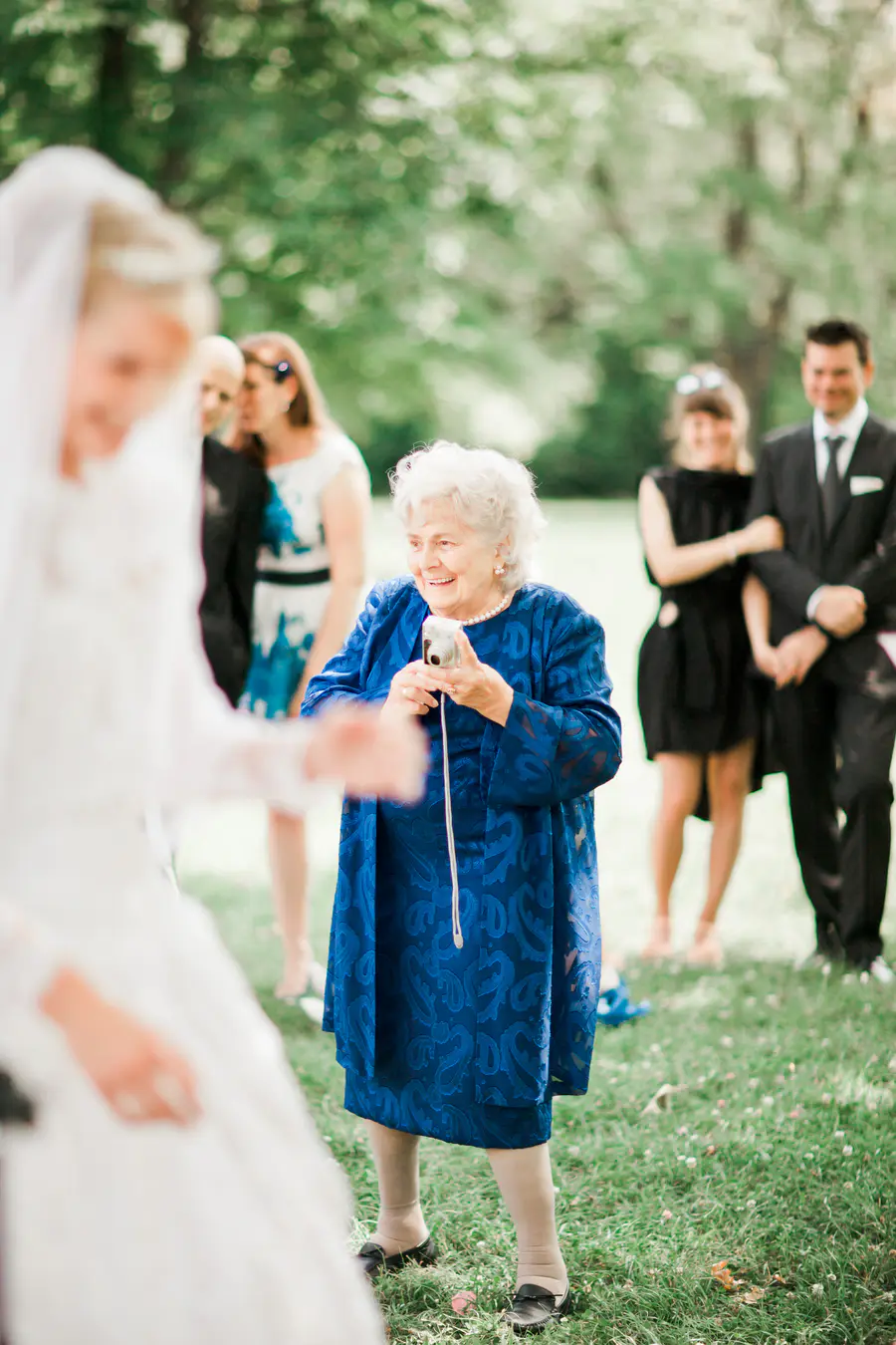 An elderly woman in a blue patterned dress smiles as she holds a silver digital camera at a wedding.