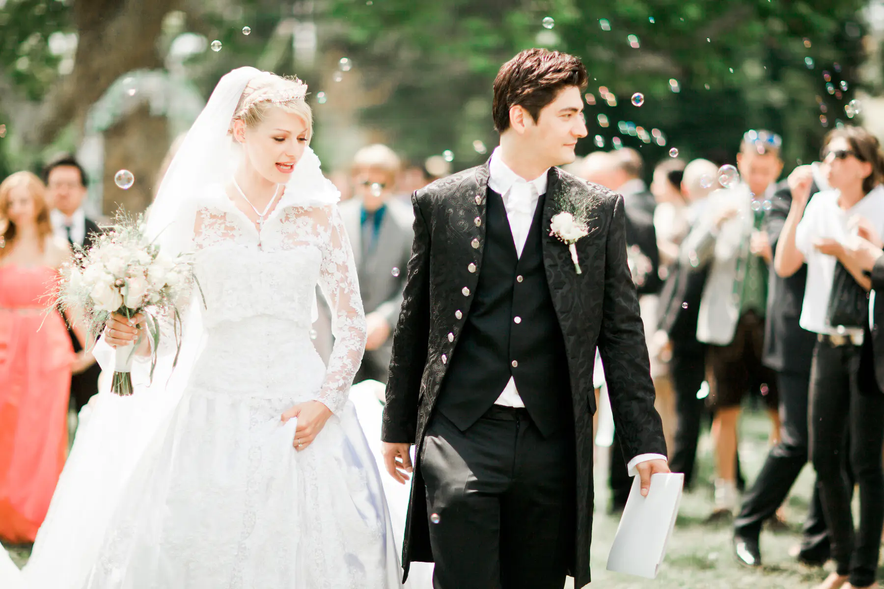 A bride and groom walk down an aisle with bubbles floating in the air. The bride wears a white lace wedding dress and veil, and carries a bouquet of white flowers. The groom is dressed in a black suit with a vest and white shirt. Other wedding guests are visible in the background.