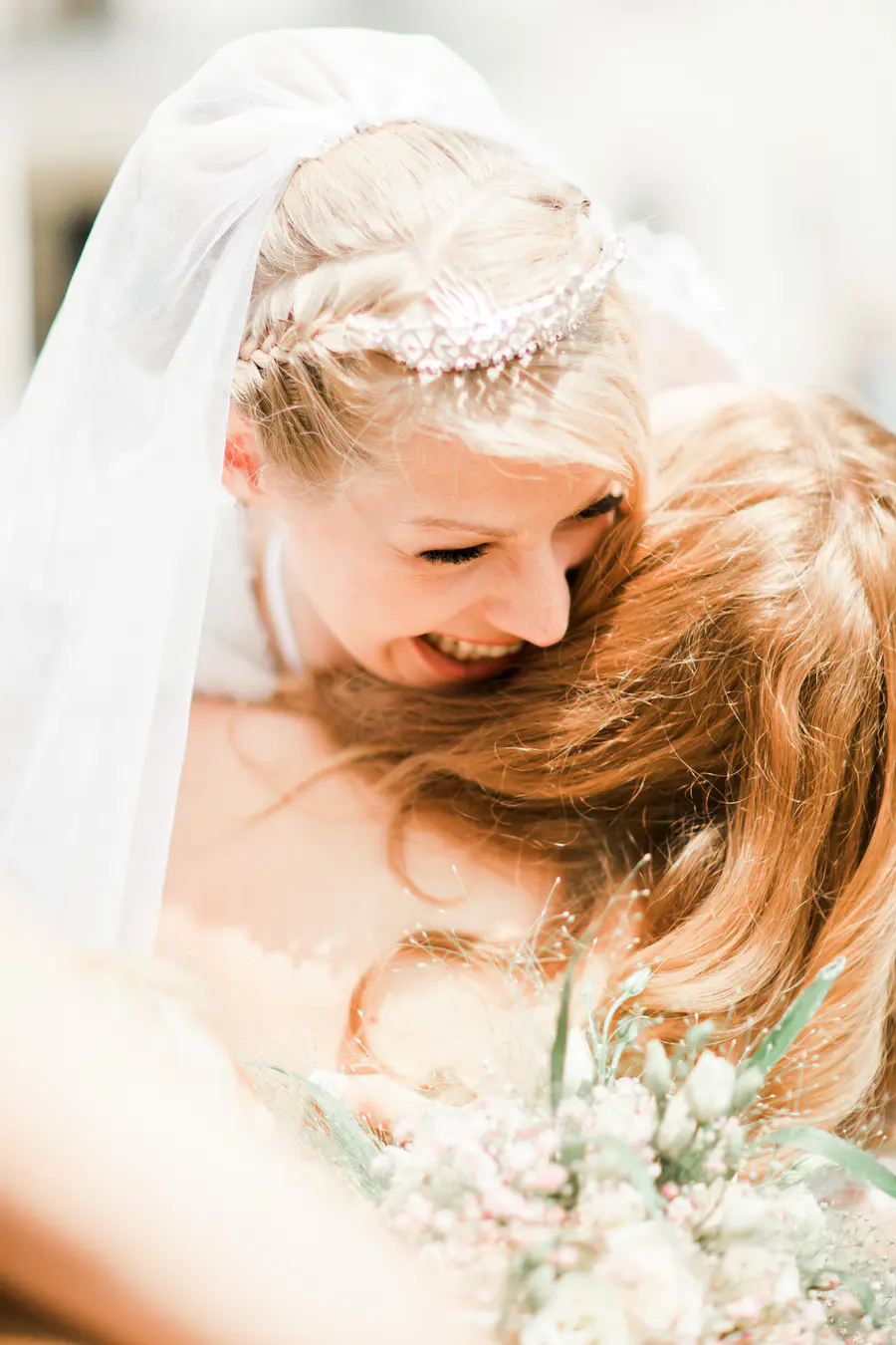 A bride with a tiara and veil embraces another person, who is mostly out of focus.