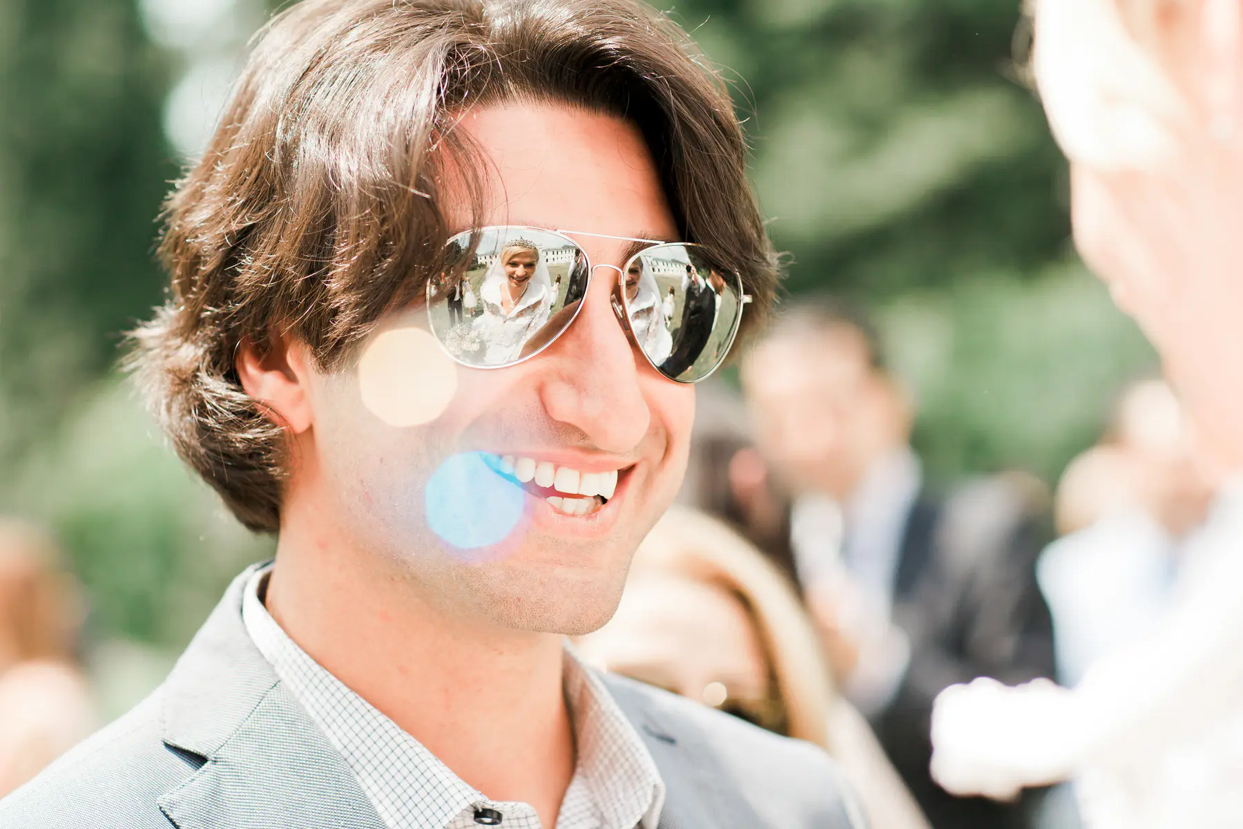A man smiles as he looks to his right, wearing aviator sunglasses that reflect a bride in a white wedding dress and a groom in a dark suit.