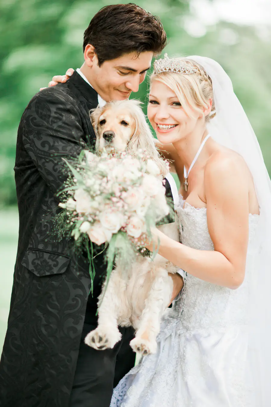 A bride and groom in wedding attire embrace a light-colored dog while holding a bouquet of rosesin details