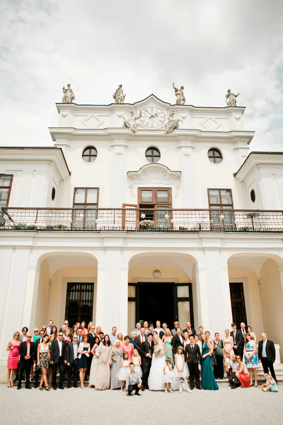 A large wedding party poses for a photo in front of a white, ornate building with a large clock on its facade.