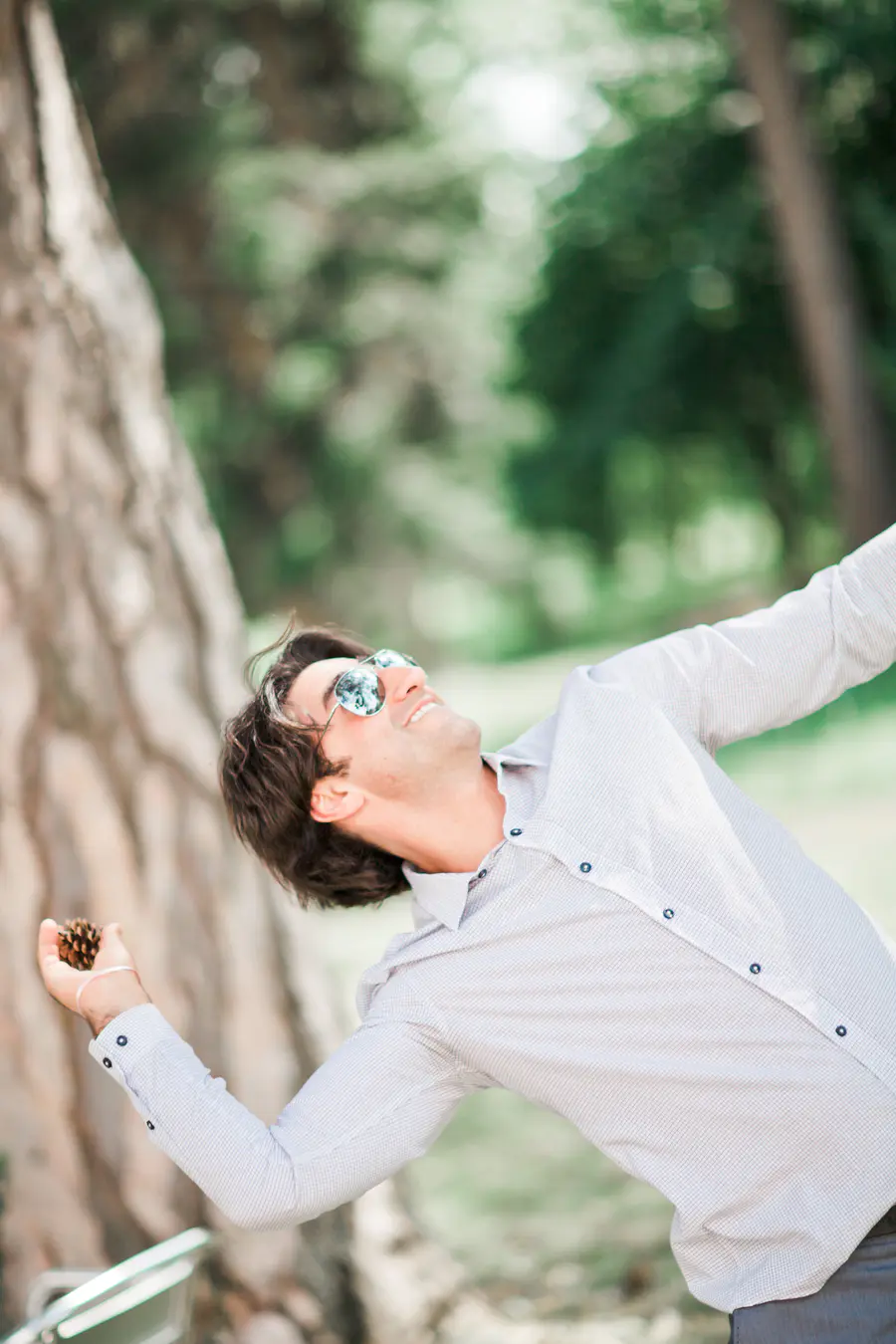 A man in sunglasses and a button-down shirt tilts his head back and smiles, holding a pinecone in his outstretched hand.
