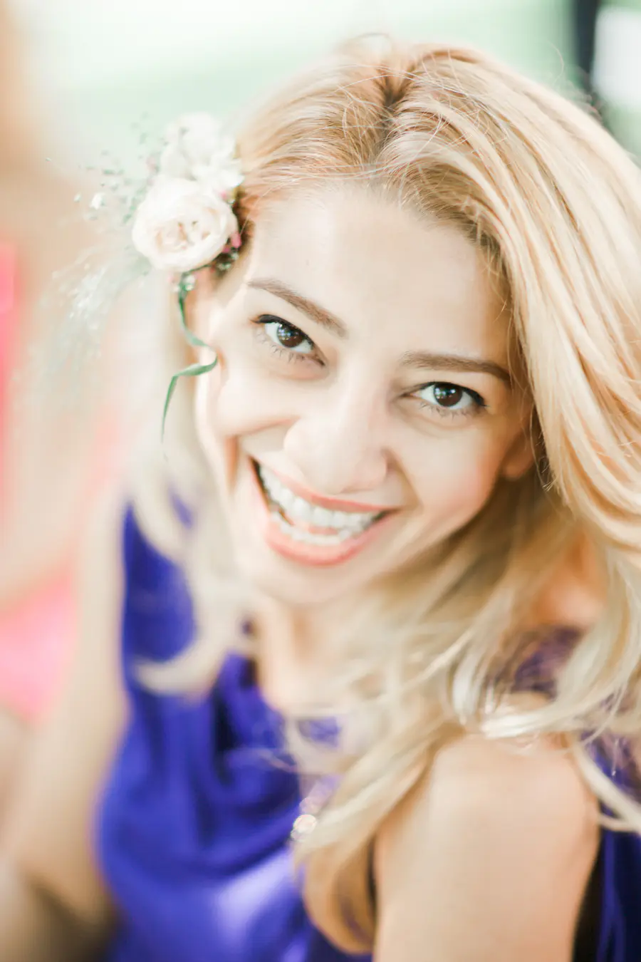 A woman with blonde hair adorned with a small cluster of white flowers smiles broadly.
