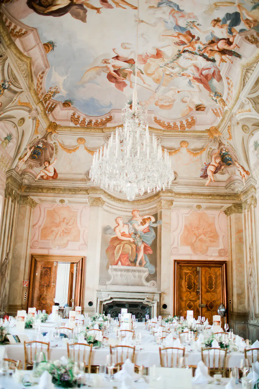 A formal dining room with a banquet set up. The room features elaborate frescoes on the ceiling and walls, a large crystal chandelier, a fireplace, and two sets of double doors.