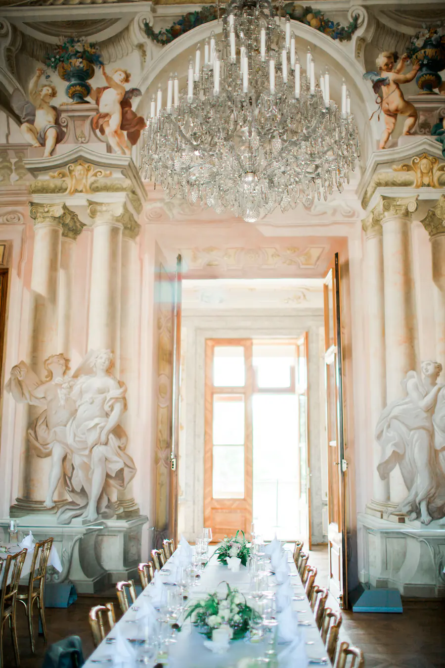 An elegant dining room in a historic building, set for a formal event, with a long table covered in a white tablecloth and set with place settings. A crystal chandelier hangs from the ornate ceiling, and sculptures of angels and cherubs adorn the walls.